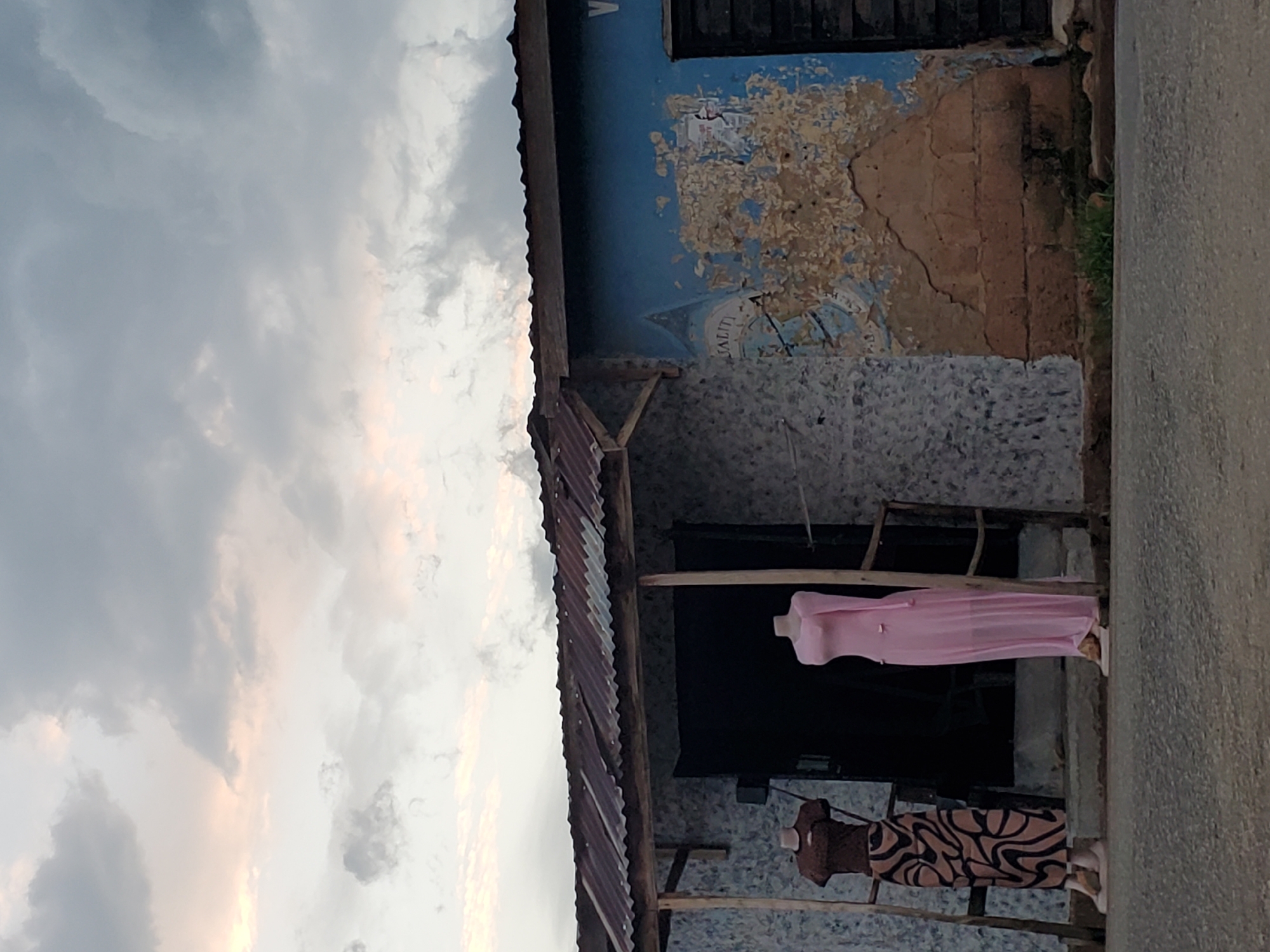 The image depicts an outdoor scene in Jos, Nigeria. The perspective is from a low angle, looking up at a structure with a corrugated metal roof and a partially exposed brick wall that has peeling blue paint and patches of discolored plaster. There are two mannequins visible, one displaying a long, flowing pink garment and the other wearing a brown top and a patterned skirt. The mannequins are placed on wooden stands, suggesting they are part of a shop display.
The sky above is filled with dramatic clouds, indicating either dawn or dusk. The lighting suggests a soft, diffused light, consistent with either early morning or late evening. The overall impression of the scene is one of a humble, possibly rural or semi-urban, commercial setting. There is a grey concrete wall on the right side of the image, and a dirt or asphalt road is visible in the foreground.
No distinct text is clearly legible in the image, although some faded lettering or logos might be present on the building's facade. The atmosphere appears quiet and still, with no people actively engaged in the scene. The focus is on the architecture, the mannequins, and the natural elements of the sky.
