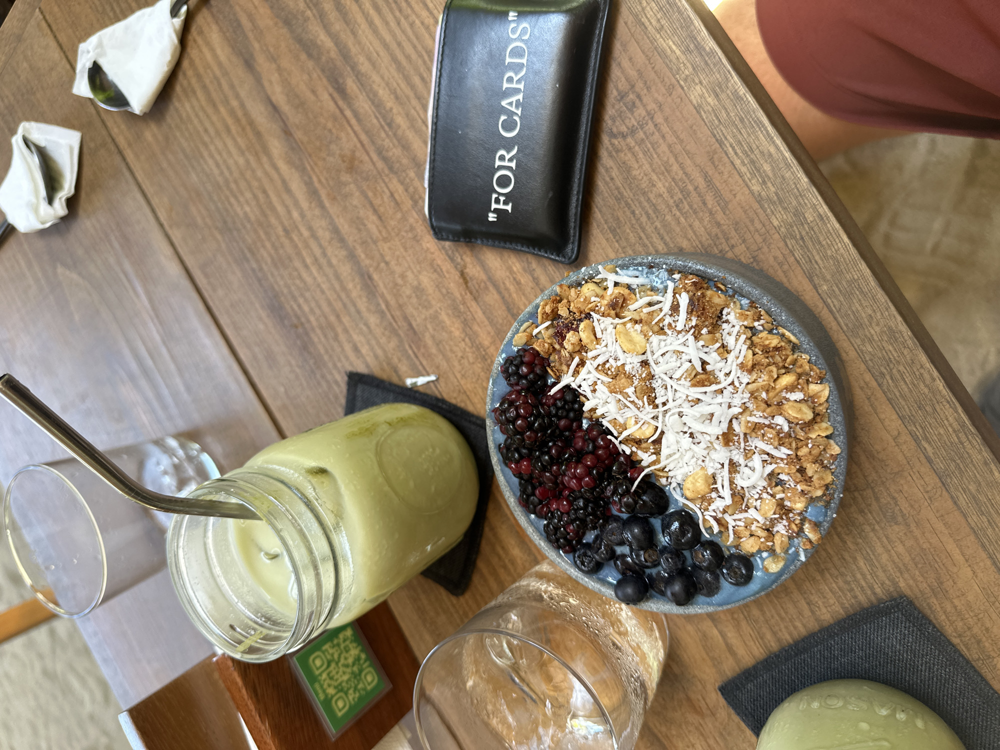 On a wooden table, potentially in a cafe or restaurant in Puerto Escondido, Mexico, sits a colorful array of food items. From top left to bottom left, there are two pieces of silverware wrapped in white napkins, a small black card holder with the words "FOR CARDS" printed on it, and a smoothie bowl with granola, coconut flakes, blackberries, and blueberries. Next to the smoothie bowl is a green drink in a glass jar with a metal straw, an empty glass, and a small green dish on a gray napkin. A person's leg is visible in the upper right corner of the image. The overall scene suggests a meal or snack being enjoyed in a casual setting.