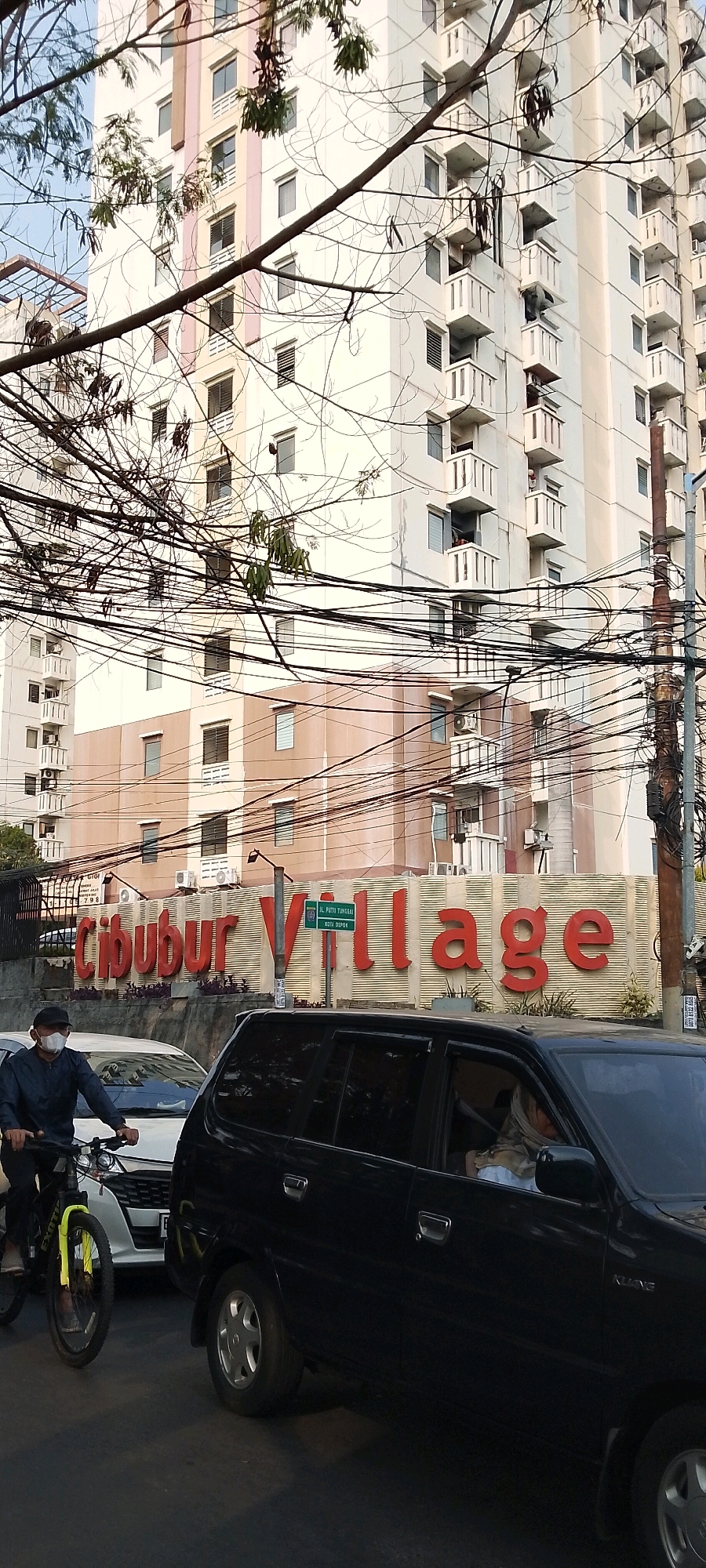 The image depicts a street scene in Depok, Indonesia, featuring a large apartment building in the background. The building is cream-colored with beige and pink accents, and features numerous balconies. A prominent sign with red lettering reading "Cibubur Village" is visible on a low wall in front of the building. A street sign indicating "Jl. Putri Tunggal Kota Depok" is also present. In the foreground, a man wearing a dark long-sleeved shirt, dark pants, a black cap, and a white face mask is riding a bicycle. The bicycle has a yellow fork. To the right of the cyclist, a black SUV is partially visible, and to the left, a white car is seen. Overhead, a dense network of black electrical wires crisscrosses the scene, partially obscuring the view of the apartment building. The lighting suggests it is daytime, possibly late afternoon or early morning, with the sun casting long shadows.