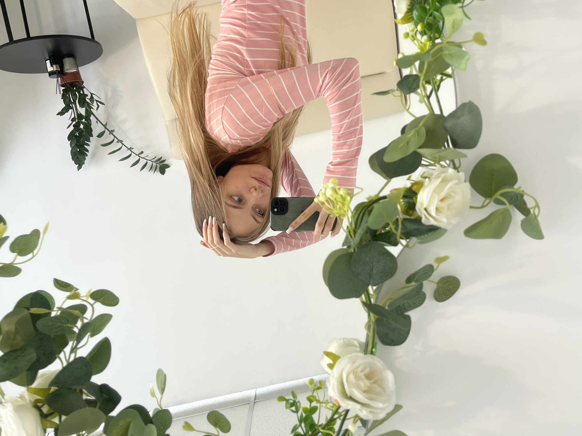 The image displays a reflection in a vertical mirror, capturing a young woman taking a selfie. The woman appears upside down in the reflection, with her head pointing towards the bottom of the frame and her body extending upwards.
The woman has long, straight blonde hair that falls towards the bottom of the frame (as it would if she were standing upright). She is wearing a long-sleeved, form-fitting top with pink and white horizontal stripes. Her face is visible, showing light-colored eyes (possibly blue or grey) looking directly towards the viewer (into the phone camera). She has light makeup and appears to have a small piercing above her upper lip. Her left hand is holding a dark green or grey smartphone, which has at least two visible camera lenses on its back. Her right hand is raised, resting near her head, with long, light-colored manicured fingernails visible, and a simple silver-toned ring on her index finger.
Behind her in the reflection, a light-colored (beige or cream) couch or sofa is visible, along with a plain white wall.
Framing the mirror's edges, particularly on the right and bottom (which would be the top and left if the image were rotated), are decorative elements. These include lush green artificial vines with various leaf shapes and several creamy white artificial roses. On the far left side of the mirror, in what would be the upper-left corner if the woman were upright, a black metal stand is visible. Suspended from this stand is a small terracotta-colored pot from which a real or artificial plant with dark green, elongated leaves hangs downwards. Small, cylindrical objects are also visible on the black stand. The overall background of the scene outside the mirror's frame is predominantly white, contributing to a bright aesthetic.