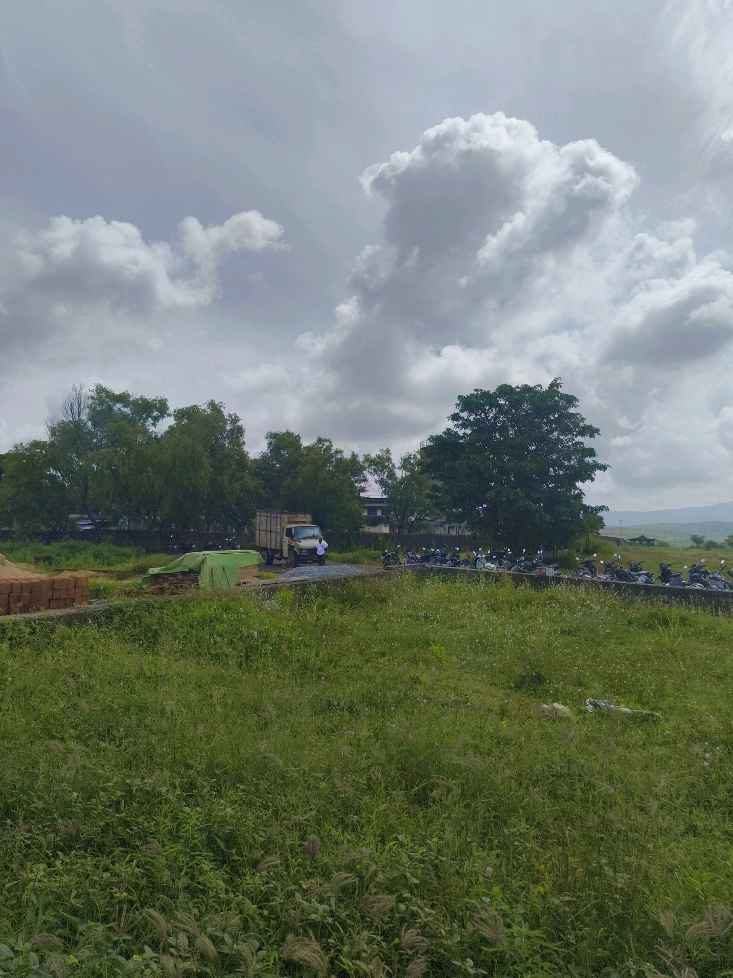 The image depicts a scene in Bhatkal, India, under a cloudy sky. In the foreground, a field of green grass is visible, with some scattered white flowers and seed heads. A low concrete barrier runs horizontally across the middle ground, separating the grassy field from a dirt and gravel area.
Behind the barrier, to the left, is a stack of reddish-brown bricks and a green tarp covering some material. Further back, a light-colored truck with a cargo bed is parked. Two men are standing near the truck, one wearing a white shirt and the other a darker shirt. To the left of the truck, a few motorcycles are parked.
Dominating the background is a dense line of trees, varying in height and foliage. Behind the trees, on the right side, there are buildings, one appearing to be a multi-story structure. In the far distance, a hazy landscape of hills can be seen. A row of motorcycles is parked along the edge of a paved area in front of the trees and buildings. The overall impression is of a rural or semi-urban setting in India.