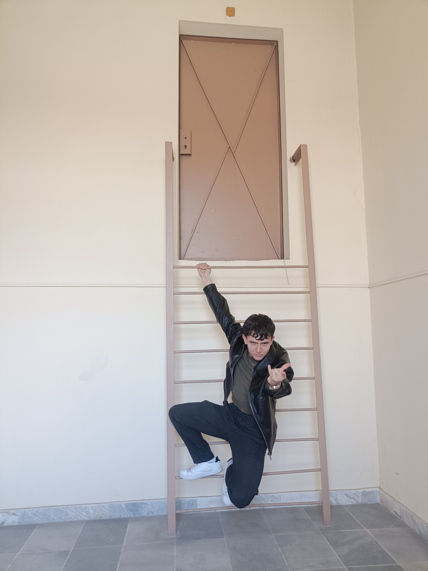 A young man is posing on a ladder in what appears to be an indoor space. He is wearing a black leather jacket, a dark green t-shirt, black pants, and white sneakers. He is gripping the top of the ladder with his left hand and has his right hand extended, showing his middle finger. The ladder is painted a light brown or beige color and is positioned in front of a door that is set high in the wall. The walls are a pale beige color, and the floor is tiled in shades of gray. The scene suggests an urban or institutional setting. The location is not discernible as Plovdiv, Bulgaria, based on the image alone.