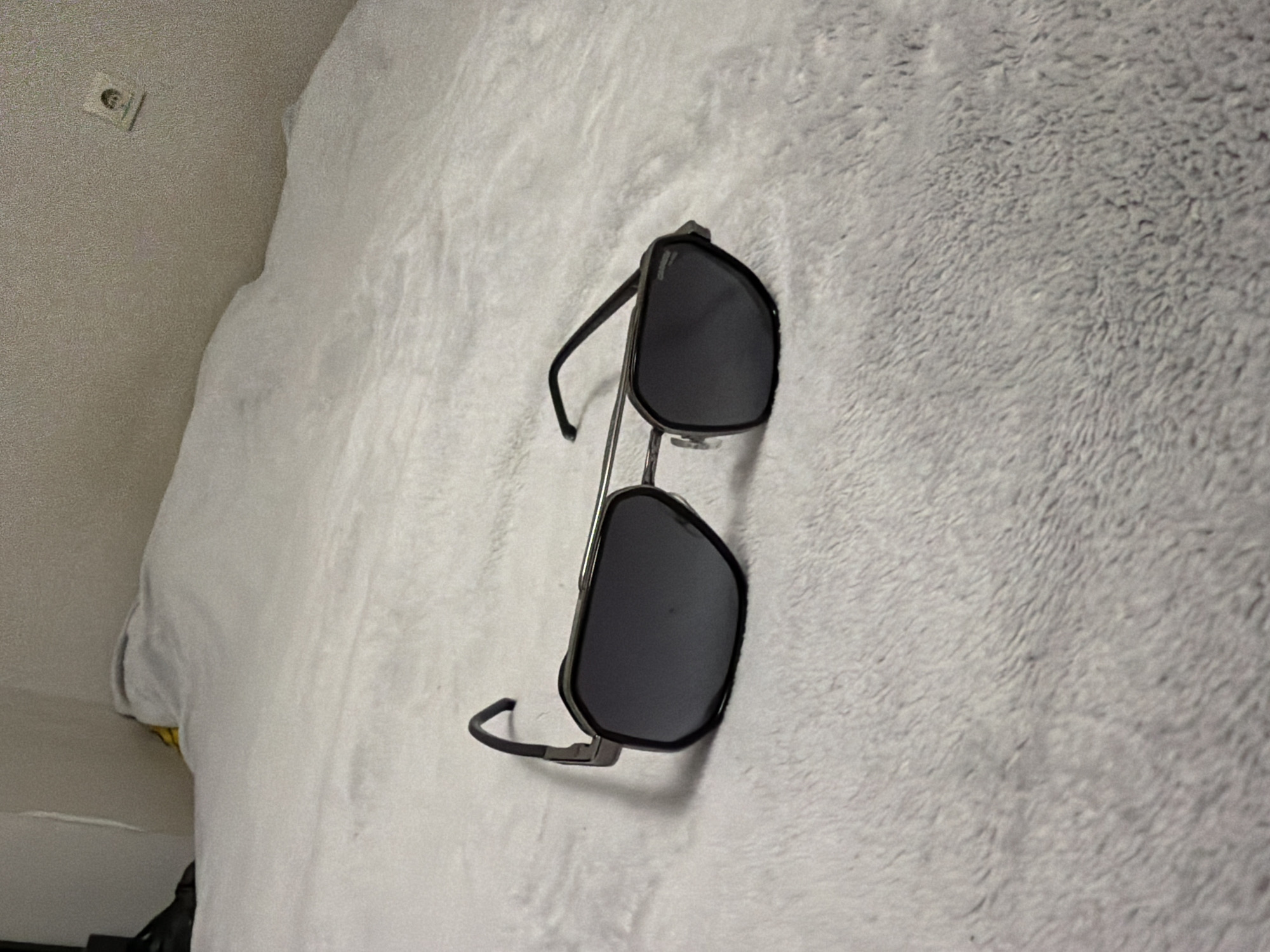 The image shows a pair of aviator-style sunglasses lying on a white, textured surface resembling a blanket or shag rug. The sunglasses have a black frame and dark lenses. In the upper left corner of the image, a section of a wall is visible with an electrical outlet. The image does not provide sufficient information to determine the location in the city of Sofia, Bulgaria.