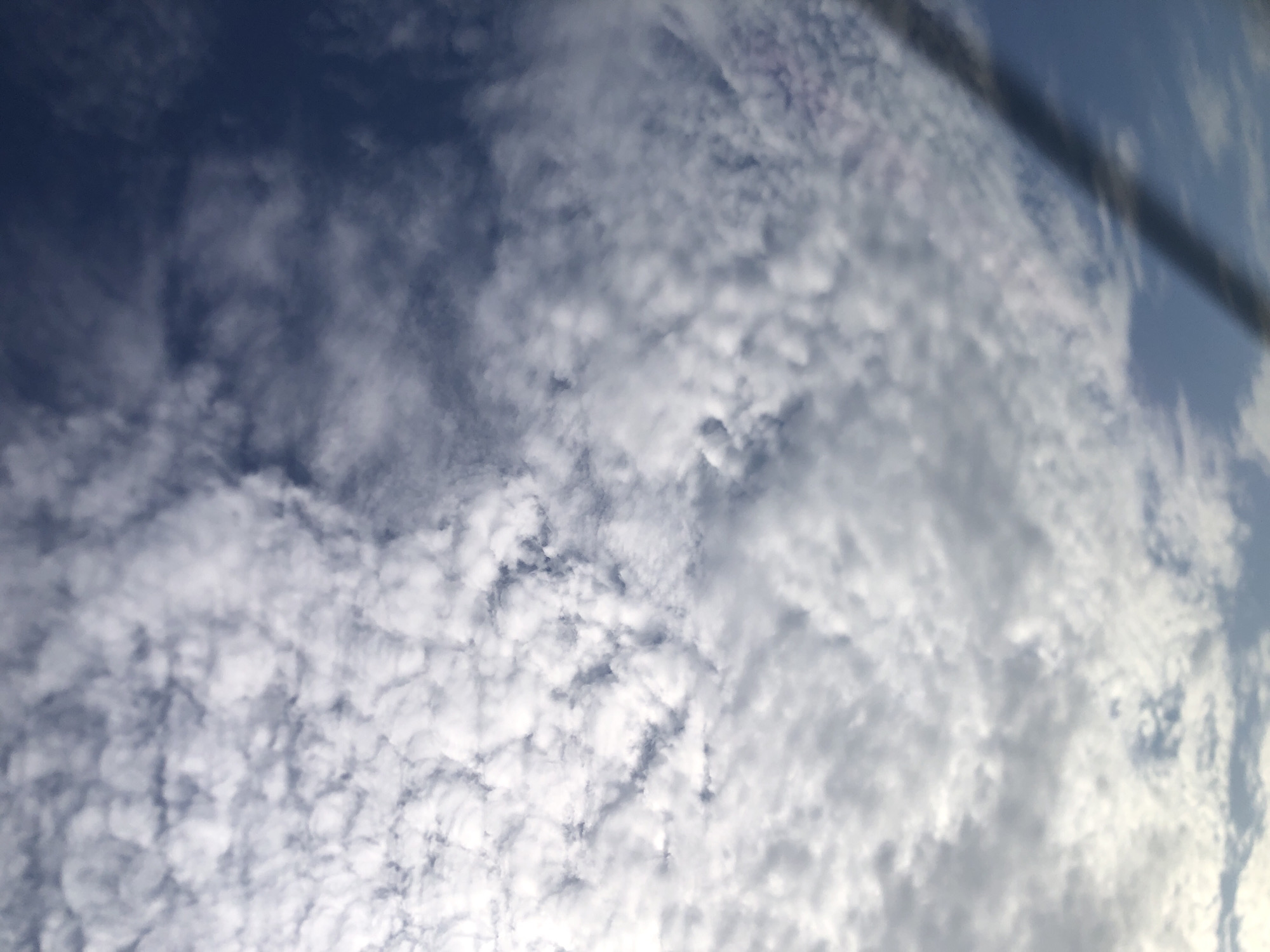 The image captures a detailed view of a cloudy sky during daylight hours. The sky is predominantly covered by white and grey clouds, exhibiting a textured, patchy, and somewhat lumpy appearance, characteristic of altocumulus or stratocumulus cloud formations. Patches of clear blue sky are visible in various areas, particularly towards the top left and right.
A prominent dark, blurred object, appearing to be a wire, cable, or a structural element, extends diagonally from the top right corner down towards the middle right of the frame, partially obscuring the clouds and sky in that region. This element suggests the photograph was taken from a specific viewpoint, possibly looking upwards from the ground or from behind an obstruction.
The lighting indicates daytime, with the clouds illuminated from above, though the sun is not directly visible. The scene is devoid of human subjects, man-made structures (apart from the blurred object), or any discernible activities. The location context for this sky view is Ukwu-Abwa, Nigeria. No text is visible in the media file.