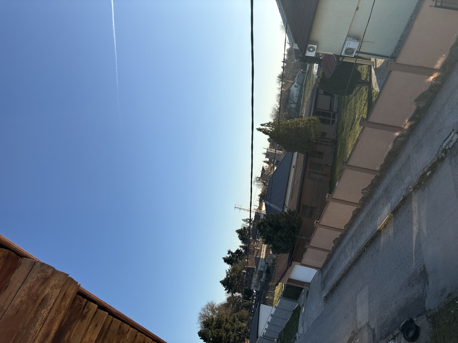 An elevated outdoor scene depicts a residential area in Stefan Karadja, Bulgaria, under a clear, bright blue sky with a single aircraft contrail visible in the upper right. A weathered wooden overhang frames the upper left corner of the image. Below, a paved street or driveway extends diagonally from the bottom left, bordered by a concrete curb. A dark, deflated tire lies on the ground at the bottom left. Properties to the right of the street are enclosed by a light brown, segmented concrete fence. Within these properties, green lawns and multiple evergreen trees are present, alongside residential buildings. One structure features wood-paneled siding and a dark tiled roof, while a cream-colored building on the far right has two external air conditioning units mounted on its facade. A dark utility cable stretches horizontally across the mid-upper portion of the frame. In the background, clusters of coniferous trees, bare deciduous trees, and a large power line tower are visible. No individuals or active vehicles are discernible within the scene.