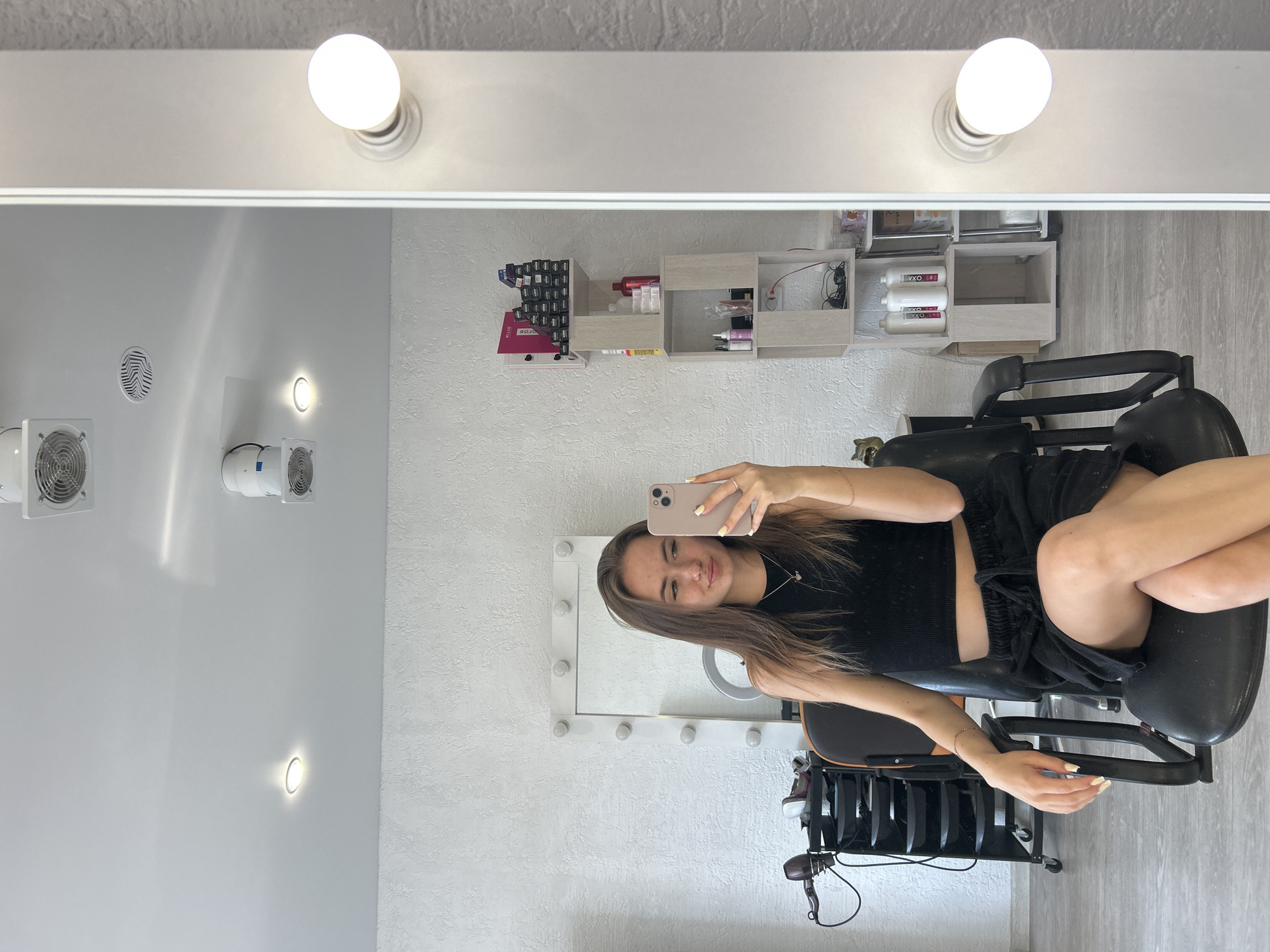 A young woman with long, brown hair, wearing a black crop top and black shorts, sits in a chair in front of a lighted mirror. She is taking a selfie with a pink smartphone. The mirror reflects a room with white walls and a gray floor. There are shelves on the wall filled with various beauty products. Above the mirror are several light bulbs, typical of a vanity setup. In the background, on the walls, there are round vents and smaller lights. A trolley with hair dryers is visible below the chair. The scene appears to be an indoor beauty salon or a personal vanity area. The lighting suggests it is daytime, but the indoor setting makes it impossible to determine the exact time of day or weather conditions. The woman has a relaxed expression on her face. No visible text is discernable in the image.