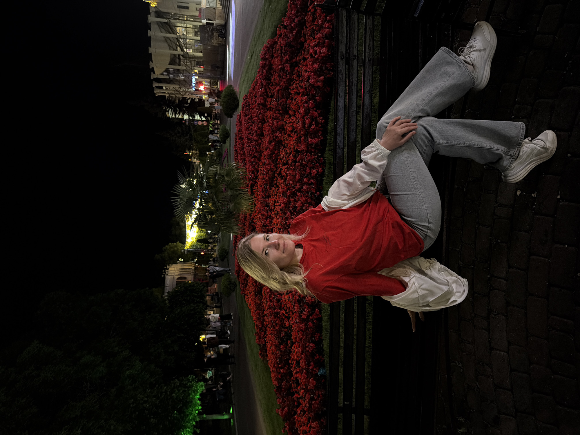 A Caucasian woman with blonde hair is sitting on a park bench at night, in front of a large flowerbed filled with red flowers. She is wearing a red long-sleeved shirt, grey jeans, and white sneakers. A white jacket is draped over her shoulders. Behind her, trees with green lights and buildings with illuminated windows are visible. The ground in front of the bench is paved with bricks. The scene appears to be set in a city park. Based on the context provided, this location is Kislovodsk, Russia.