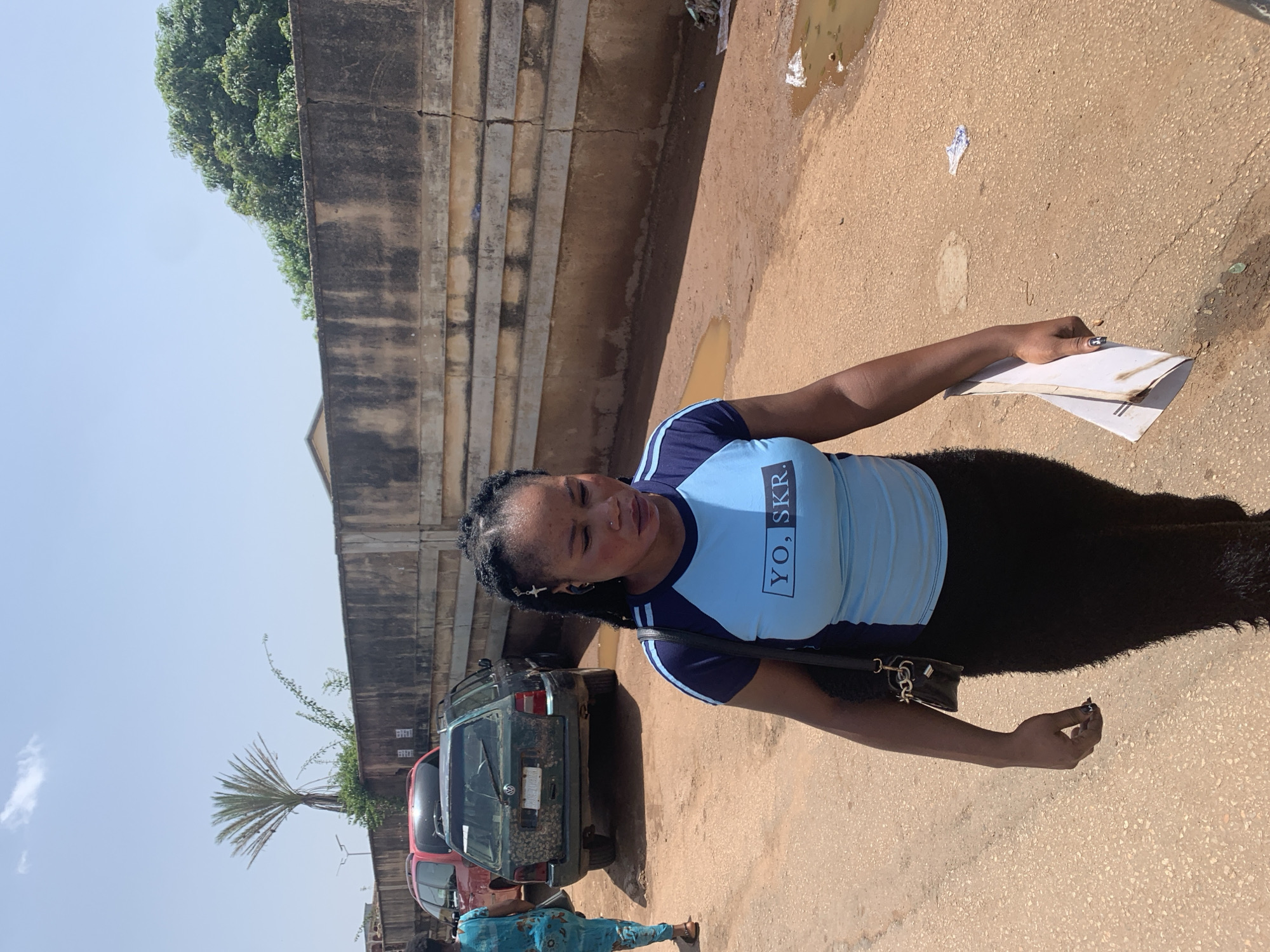 A black woman with braided hair and a light blue t-shirt with "YO. SKR." printed on it walks down a dirt road, holding a white folder. She is wearing a black skirt. She has a small black purse on her shoulder. Behind her, a green station wagon and a red SUV are parked. The road is dusty, and there are puddles of water on the ground. A concrete wall runs along the side of the road, with some greenery growing on top. The sky is clear and blue, with a few clouds. The scene appears to be in the city of Jos, Nigeria.