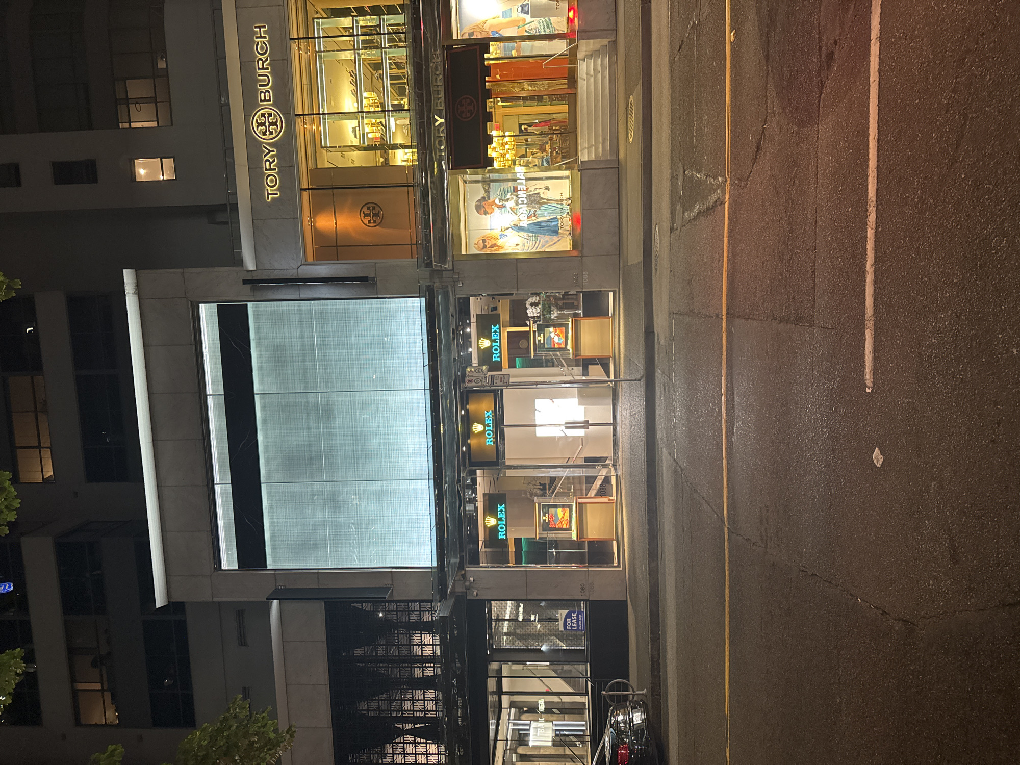 This is a nighttime street-level photograph of a commercial building facade in Vancouver, Canada. The building features a prominent, large, illuminated digital display screen across its upper level. Below this screen, there are three distinct storefronts. The central storefront is a Rolex store, clearly marked with its iconic logo and name on illuminated signs. The interior of the Rolex store is visible, showcasing display cases with watches. To the right of the Rolex store is a Tory Burch store, also indicated by its logo and name, with a brightly lit interior displaying merchandise. To the left of the Rolex store is another storefront, partially obscured by foliage in the upper left corner of the image, which appears to be Balenciaga, with a "FOR LEASE" sign visible in the window. The street in front of the building is paved and appears wet, reflecting the lights from the storefronts. A black car is parked in front of the Balenciaga store, and a bicycle rack is also present. The environment suggests a busy urban commercial district.