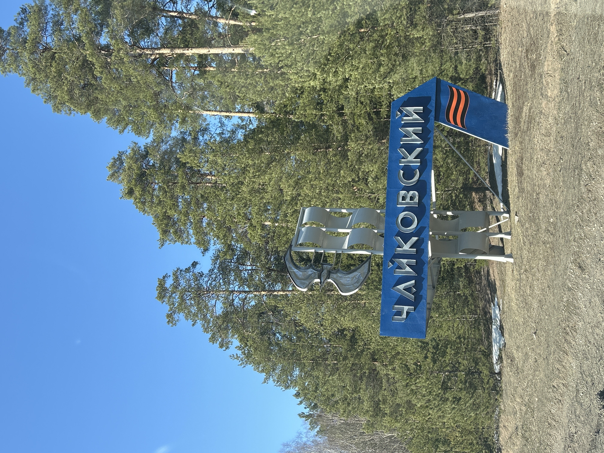 A roadside sign indicates entry to Chaikovsky, Russia. The sign is blue with silver Cyrillic lettering. A stylized metal sculpture sits on the left side of the sign. An orange and black ribbon emblem is on the top right. The sign is surrounded by a forest of tall pine trees. A clear, blue sky is visible. The ground cover appears to be dry grass and dirt.