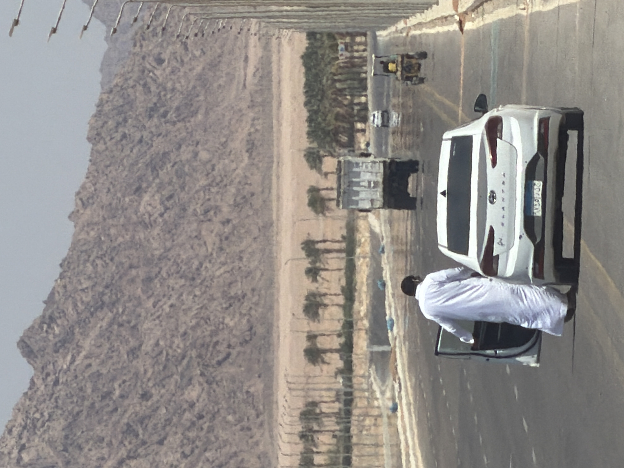 A man in a white thobe exits a white Hyundai Elantra on a wet asphalt road in an arid environment. The car's driver's side door is open. In the background, a flatbed truck drives away from the viewer, followed by a small tractor with a person driving it, and then a white sedan. Palm trees and more sparse vegetation are visible beyond the road, and rocky, barren mountains form the distant horizon. The sky is clear and pale blue. The scene appears to be along a highway in Egypt, given the license plate on the Hyundai.