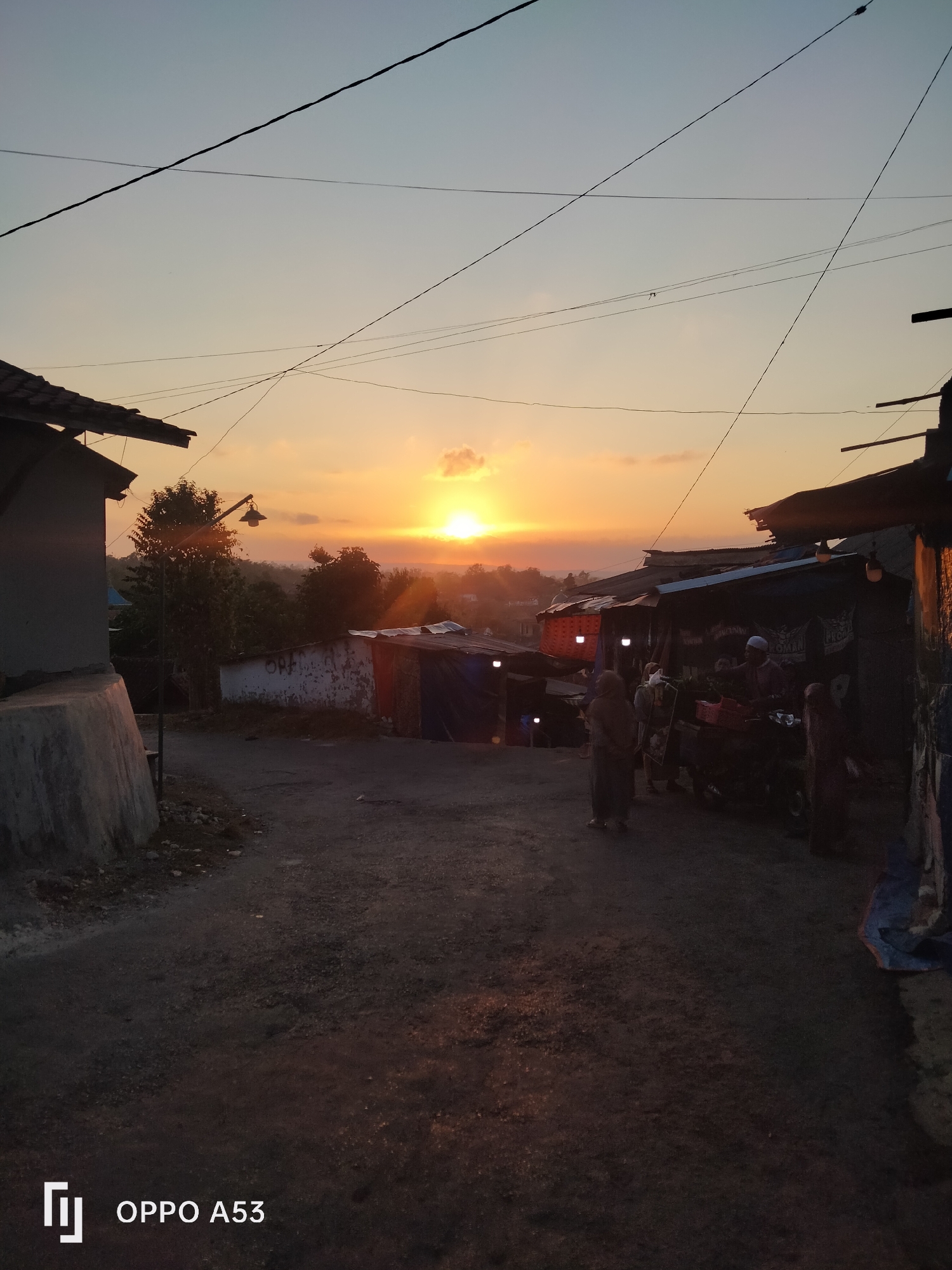 This is a vertical, medium-shot, eye-level photograph of a street scene in what appears to be a rural or semi-urban area of Badung, Indonesia, during sunset. The sun is setting on the horizon, casting a warm orange and yellow glow across the sky, with scattered clouds visible. The foreground features a dirt road that curves gently. On the left, a concrete structure, possibly part of a building or a retaining wall, occupies the immediate left edge. Further back on the left, there is a building with a tiled roof and some trees.
The right side of the street is lined with makeshift stalls or small shops, some covered with corrugated metal and tarps. These structures appear to be selling goods, as evidenced by the presence of people and what look like items for sale. In the middle ground, several individuals are visible. Two people are standing near the stalls, one appearing to be a vendor at a stall with produce. Another person is walking away from the camera down the road. The lighting suggests it is dusk, with artificial lights from the stalls adding to the illumination. Power lines are visible crisscrossing the sky. The overall impression is one of a typical evening scene in a local marketplace in Indonesia. The bottom left corner of the image contains the watermark "OPPO A53."
