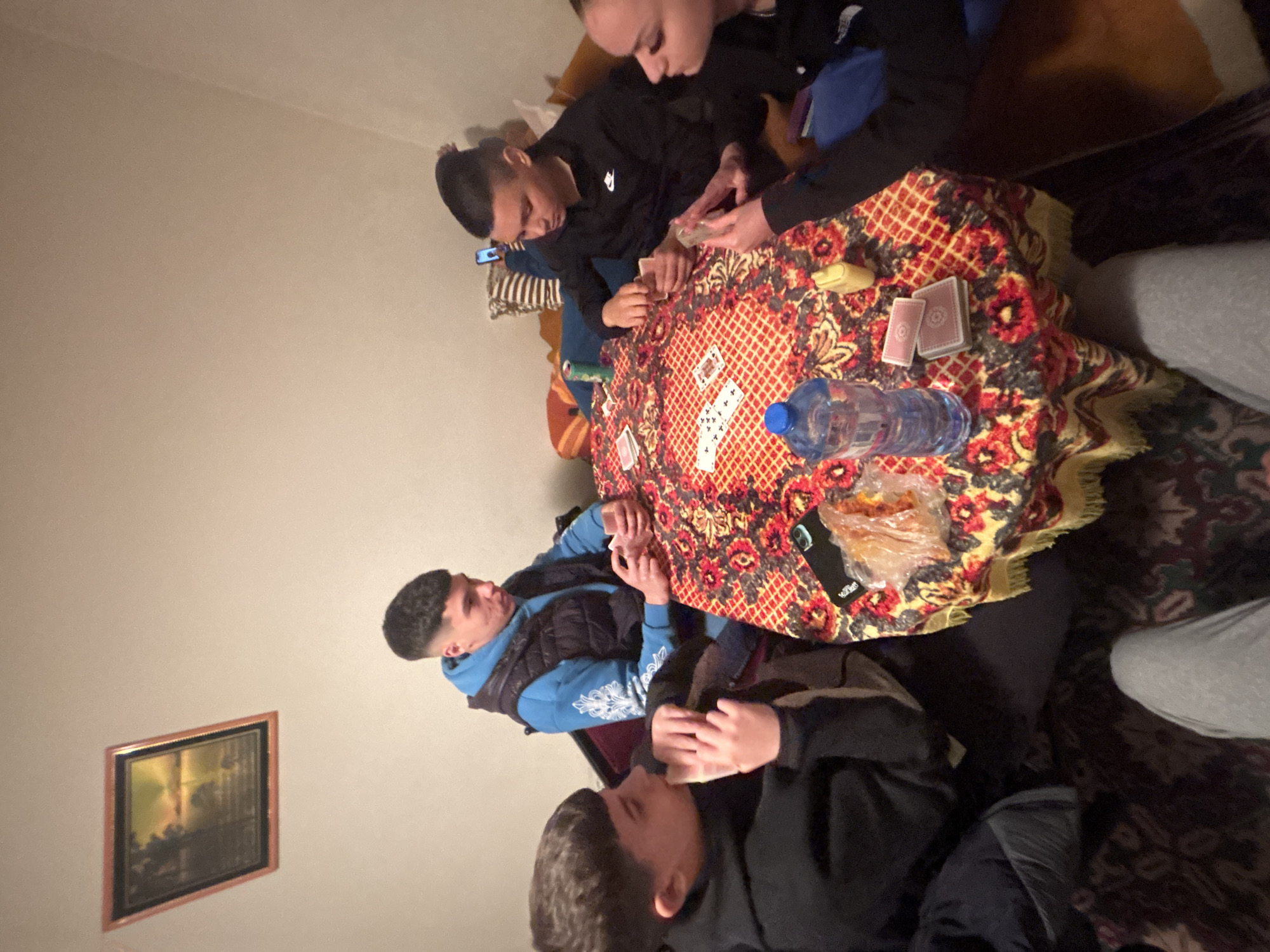 Four young men are seated around a round table covered with a patterned tablecloth. Playing cards are scattered across the table, along with a water bottle, a bar of soap, a cell phone, and a plastic bag containing food. The men appear to be engaged in a card game. The room has beige walls and a framed painting hangs on the wall. No information to suggest location is in Goritsa, Bulgaria.