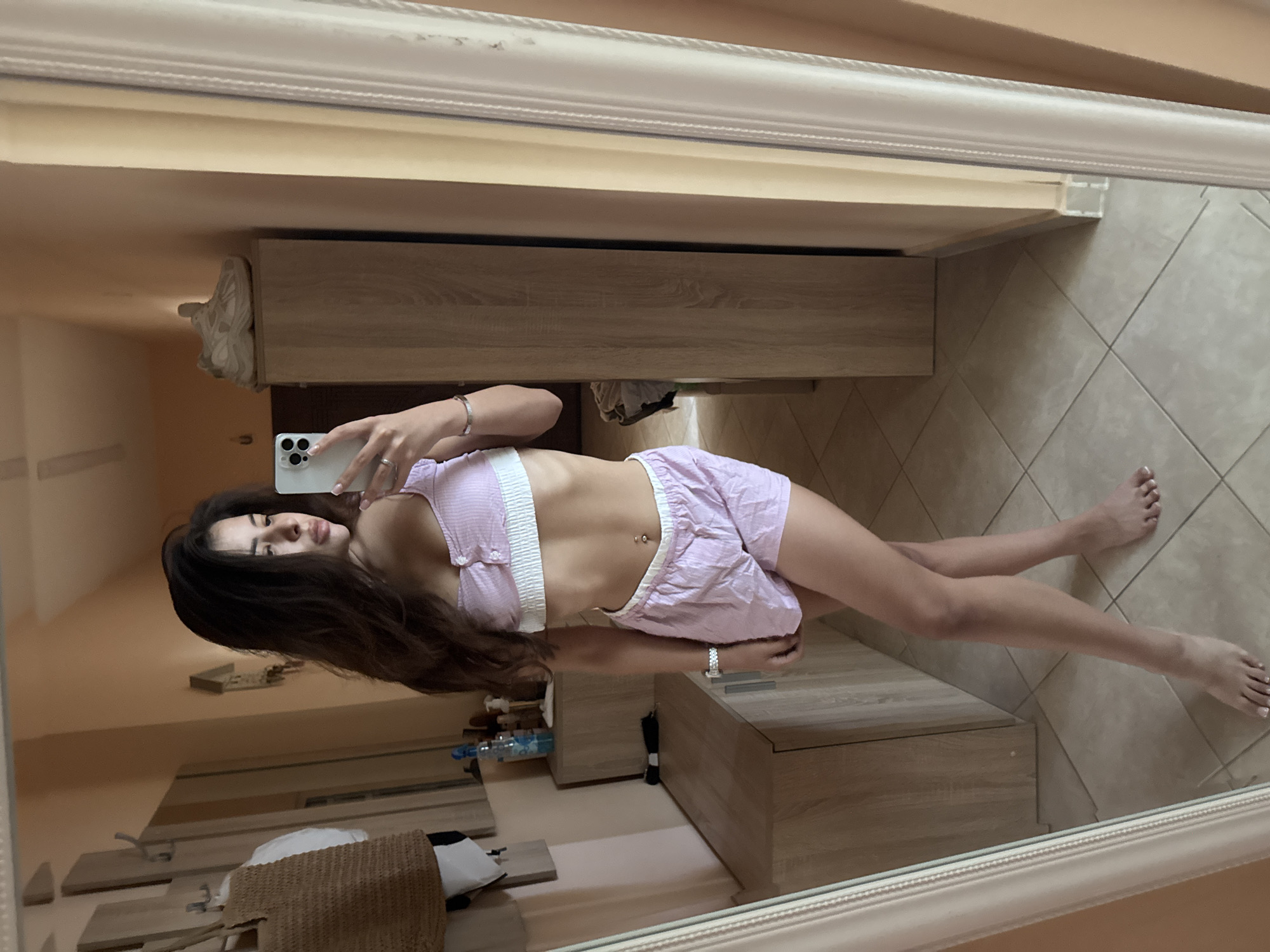 A young woman is taking a mirror selfie in what appears to be a bedroom or dressing area. She is wearing a pink and white striped crop top and matching shorts. Her long, dark hair is down, framing her face. She is holding a white smartphone in her right hand. Her left arm is bent, and her hand is resting on her hip. The woman has a flat stomach with a navel piercing. Her feet are bare and are visible in the reflection.
The background of the mirror shows a room with tiled flooring and wooden furniture, including a dresser or wardrobe. A woven bag is also visible in the reflection. The overall setting suggests an indoor, possibly residential, environment. The lighting appears to be natural, possibly from a window, creating a soft illumination. There are no discernible signs of text within the image. The image is captured in portrait orientation.
