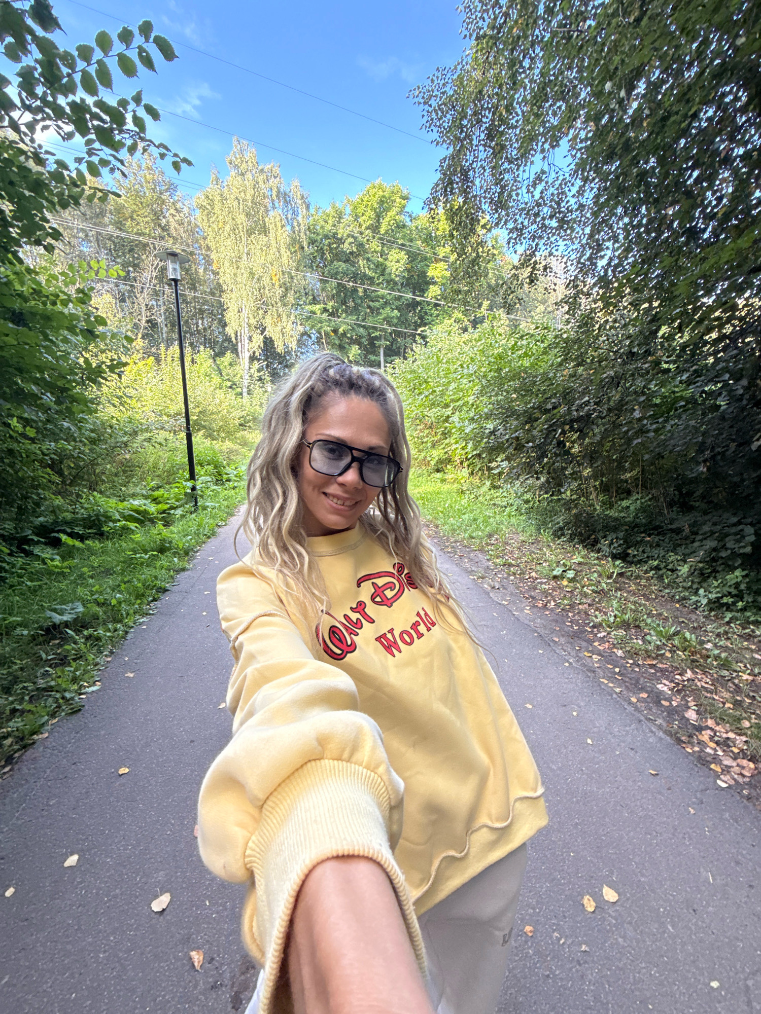 A woman is taking a selfie while walking on a paved path through a park. She is wearing a yellow sweatshirt with "Disney World" printed in red letters on the front and large, dark sunglasses. Her blonde hair is styled in loose waves and pulled back into a ponytail. The path is lined with lush green trees and foliage. A lamppost is visible in the background. The sun is shining, indicating it is daytime. The overall mood is cheerful and relaxed. There are fallen leaves on the path, suggesting the season might be autumn.