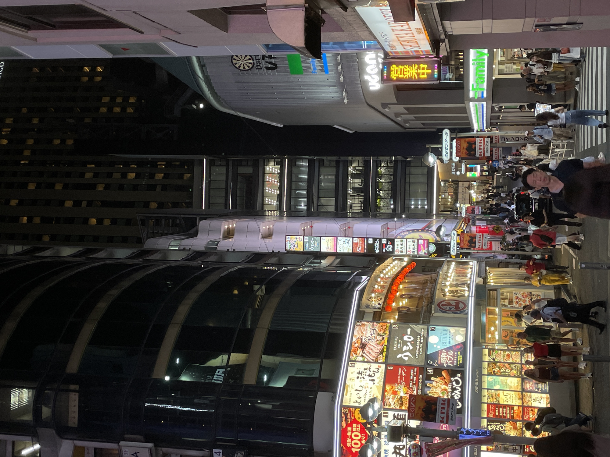 This is a nighttime street scene in Shibuya, Japan. The street is lined with tall, modern buildings, many of which have illuminated signs and advertisements. People are walking along the sidewalk and crossing the street. Some of the businesses visible include a FamilyMart convenience store, and restaurants with Japanese characters on their signs, featuring images of food such as yakiniku. The overall atmosphere is that of a bustling urban environment at night.