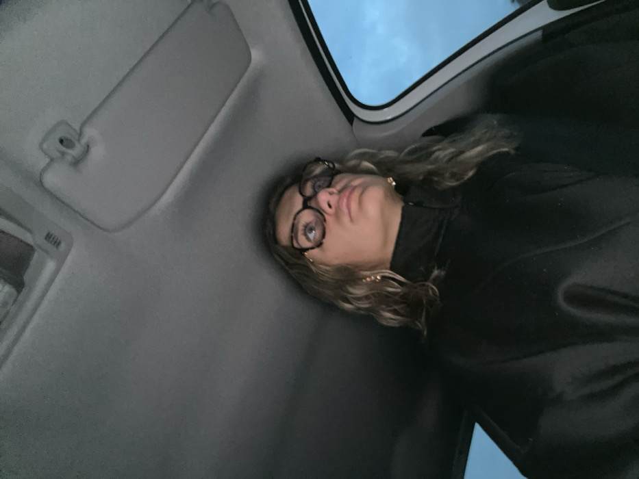A woman with blonde, wavy hair and glasses is in a car. She is wearing a black top. The car's interior is grey. The sun visor is down on the driver's side. The car window shows a cloudy sky.