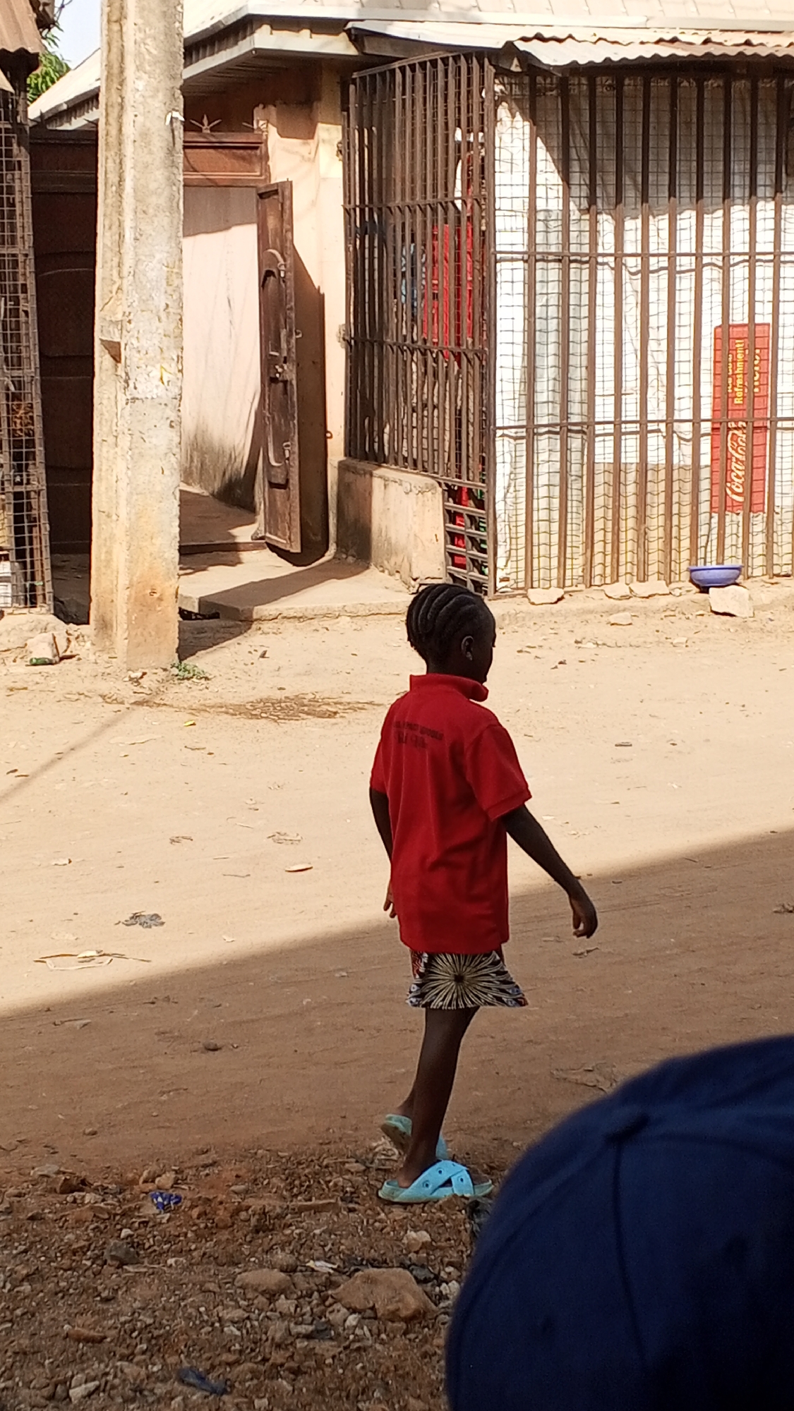 A young girl with braided hair, wearing a red shirt and patterned skirt, walks on a dirt road in Sabon Tasha, Nigeria. She wears light blue sandals. Buildings with metal bars over windows and doors flank the road. A partially visible figure wearing a dark blue hat is in the foreground.
