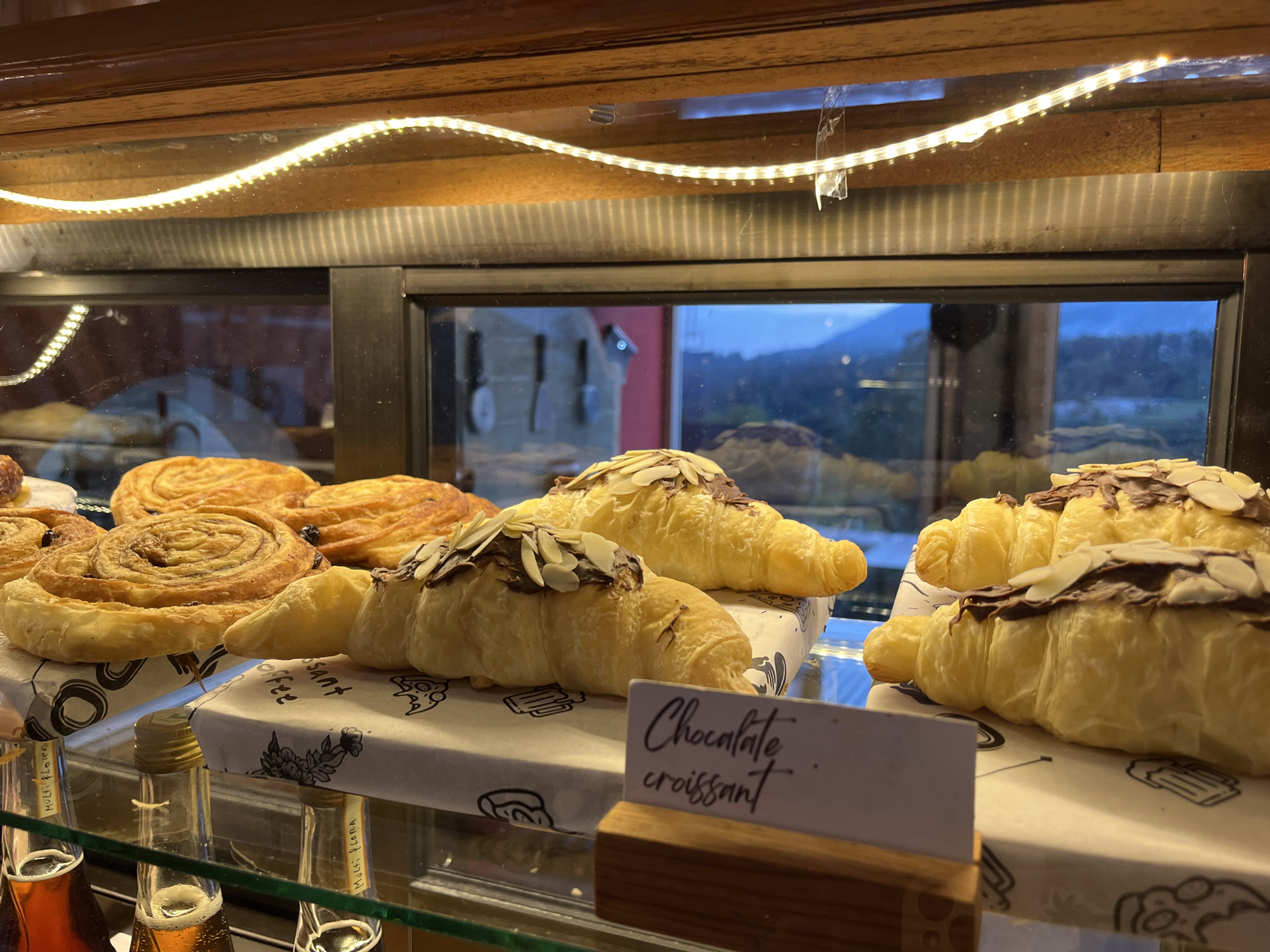 A display case in Baturiti, Indonesia, showcases pastries. A row of danishes is on the left, followed by chocolate croissants topped with sliced almonds. A sign reads "Chocolate Croissant". Below, partially visible, are bottles of what appear to be syrup. In the background, seen through the glass, is a view of mountains and buildings, consistent with the Indonesian landscape. A warm light strip illuminates the pastries from above.