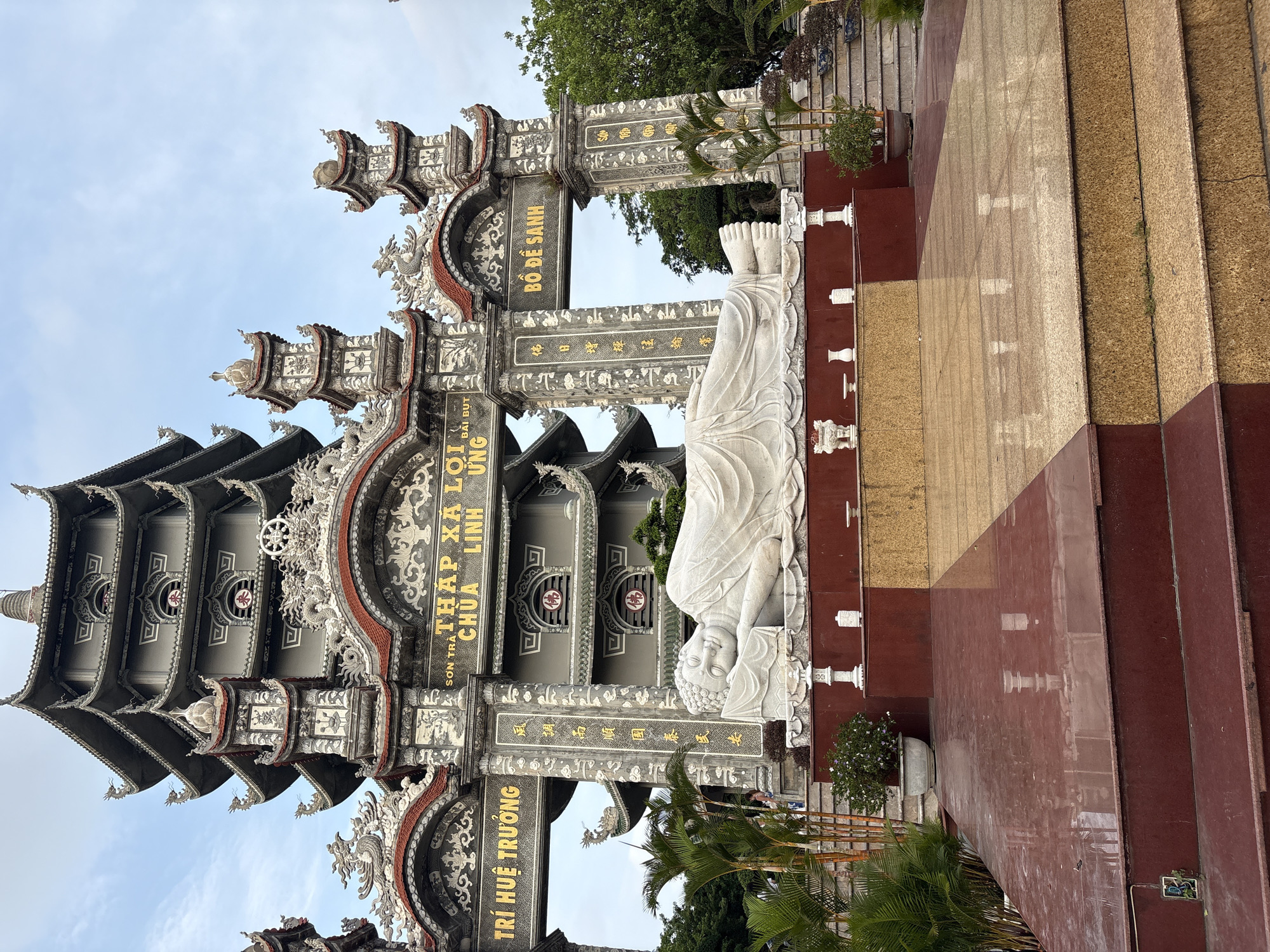 This is a street-level, medium shot of a Buddhist temple in Đà Nẵng, Vietnam. The temple architecture features multiple layered roofs with intricate dragon sculptures adorning the edges. Golden characters in vertical columns stand out against the light gray background of the temple structures. A white marble statue of a reclining Buddha is positioned near the entrance. In the foreground, stairs with dark red and tan colored stones lead up to the temple complex, and palm trees and greenery add a natural element to the scene.
