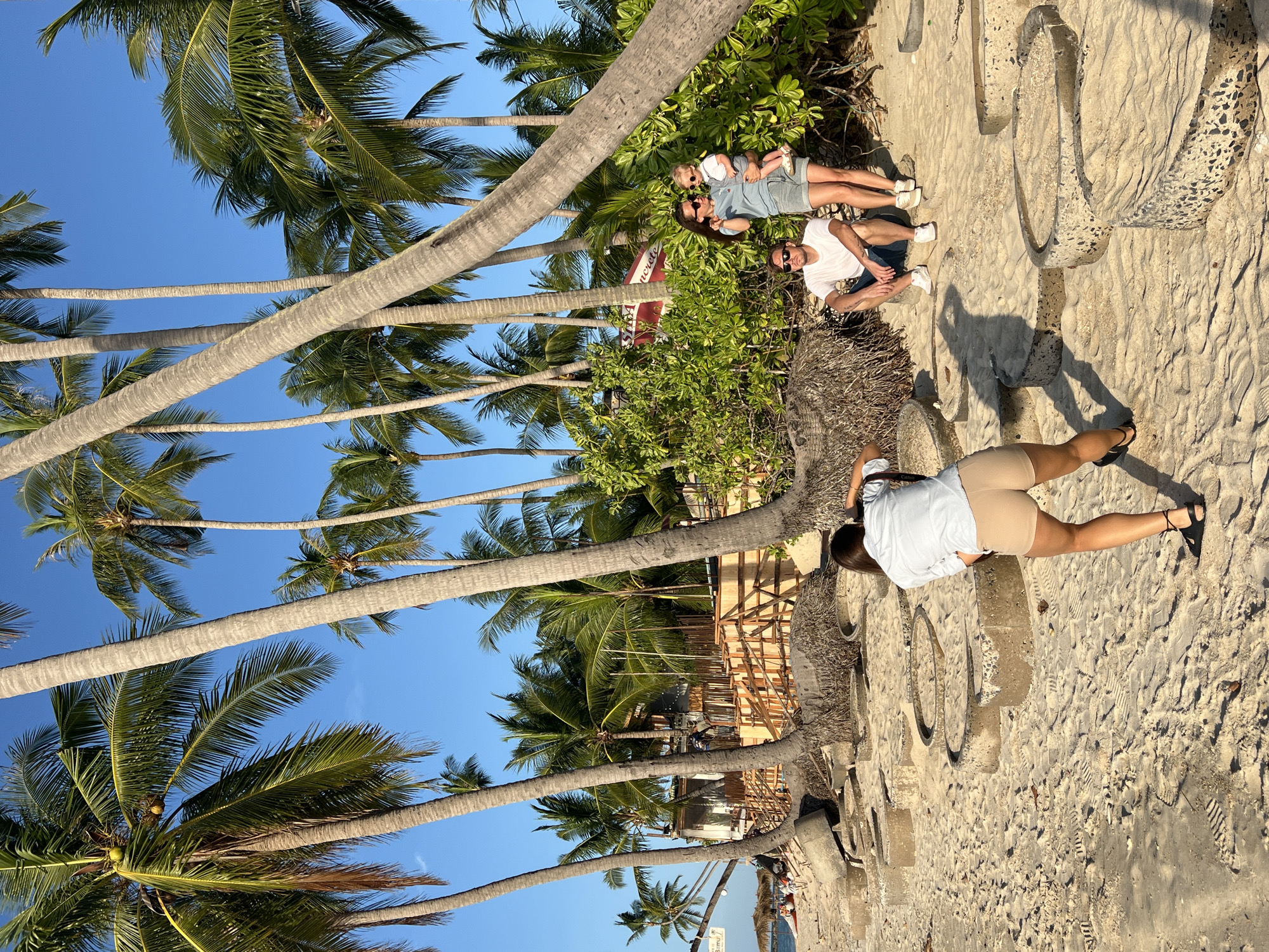 The image depicts a beach scene in Baan Tai, Thailand. Palm trees dominate the left side of the frame against a clear blue sky. A family is positioned to the right of the trees: a woman in sunglasses holding a young child, and a man seated next to them. Further to the right, a woman is kneeling on the sand, appearing to take a photo. The sand is marked with footprints, and there are concrete circles embedded in the sand near the kneeling woman. In the background, there is evidence of construction and some beachside structures.