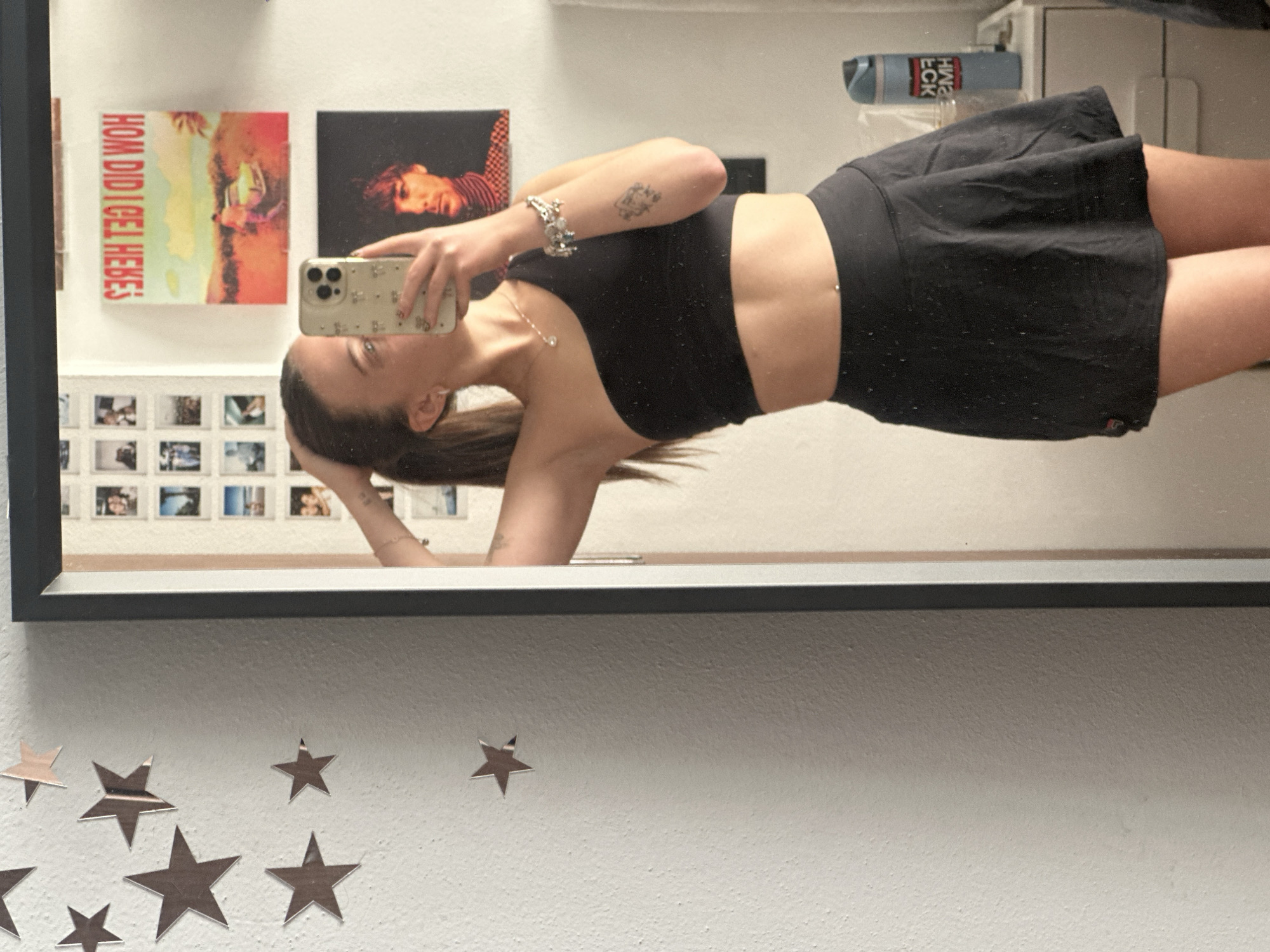 A person is taking a mirror selfie. The person is wearing a black sports bra and a black skirt. The background includes a white wall with star-shaped decorations, pictures, and what appear to be album covers. Huixquilucan de Degollado, Mexico is not discernible from this image.