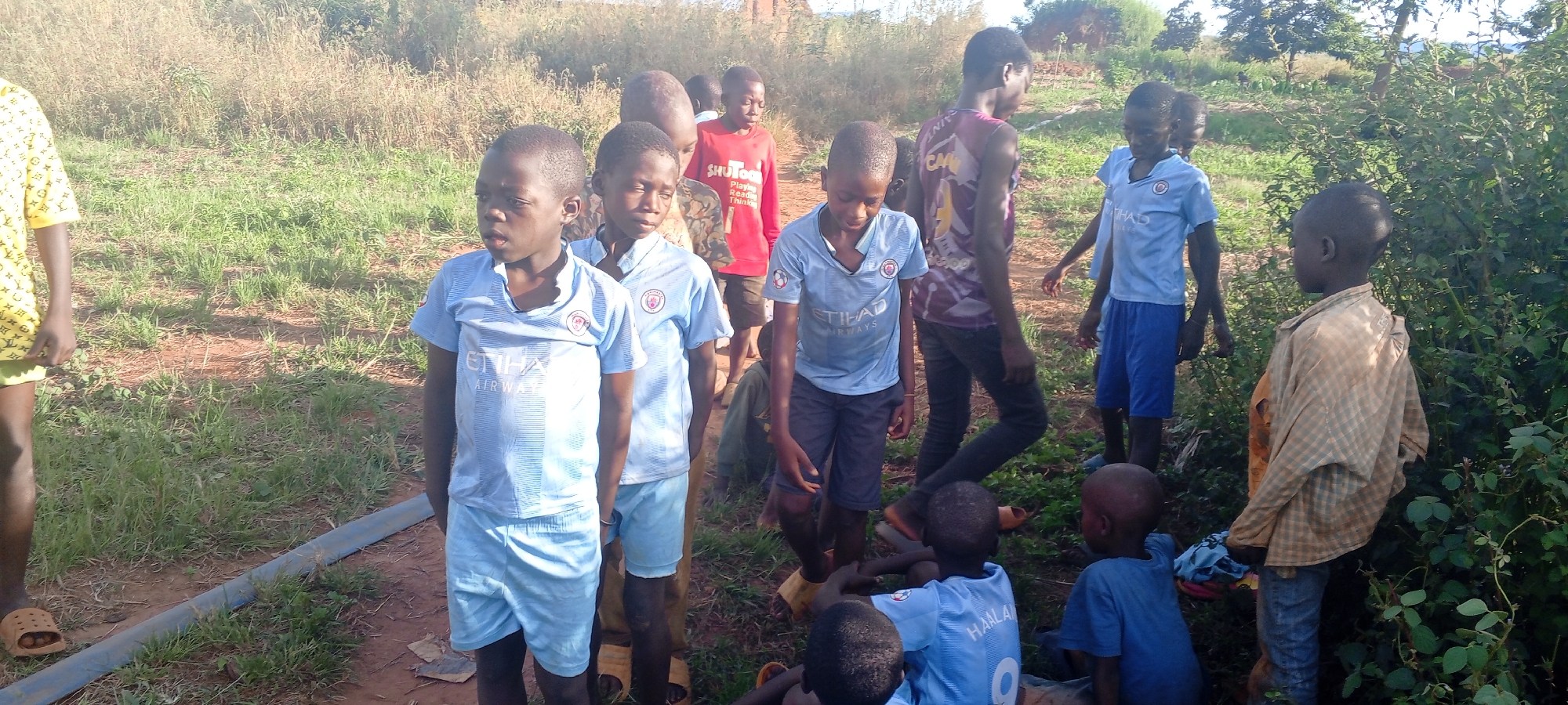 The image captures a group of approximately 14 children and young teenagers gathered outdoors in Ngalula, Democratic Republic of the Congo. The setting appears to be a rural or semi-rural area, characterized by reddish-brown dirt paths, scattered patches of green grass, and wild bushes and trees forming a natural backdrop. The bright, natural lighting indicates it is daytime, likely late afternoon, with clear weather.
Most of the children are standing, appearing to be observing their surroundings or simply congregating. Their expressions are generally neutral or watchful. Several boys are wearing light blue football jerseys; these feature "ETIHAD AIRWAYS" prominently printed on the front along with the Manchester City FC crest. One boy, seen from behind, has "HAALAND 9" visible on the back of his light blue jersey. Other clothing includes a red t-shirt with "SHUTOO" and "Playing Reading Thinking" displayed on its front, and a yellow t-shirt with a repeating pattern worn by an individual on the far left. A boy on the right is wearing a light brown and white plaid-patterned shirt.
In the lower foreground, a dark pipe or hose runs across the dirt ground. Two younger children are visible sitting or squatting towards the lower right. The overall scene depicts an informal gathering of children in an undeveloped outdoor environment.