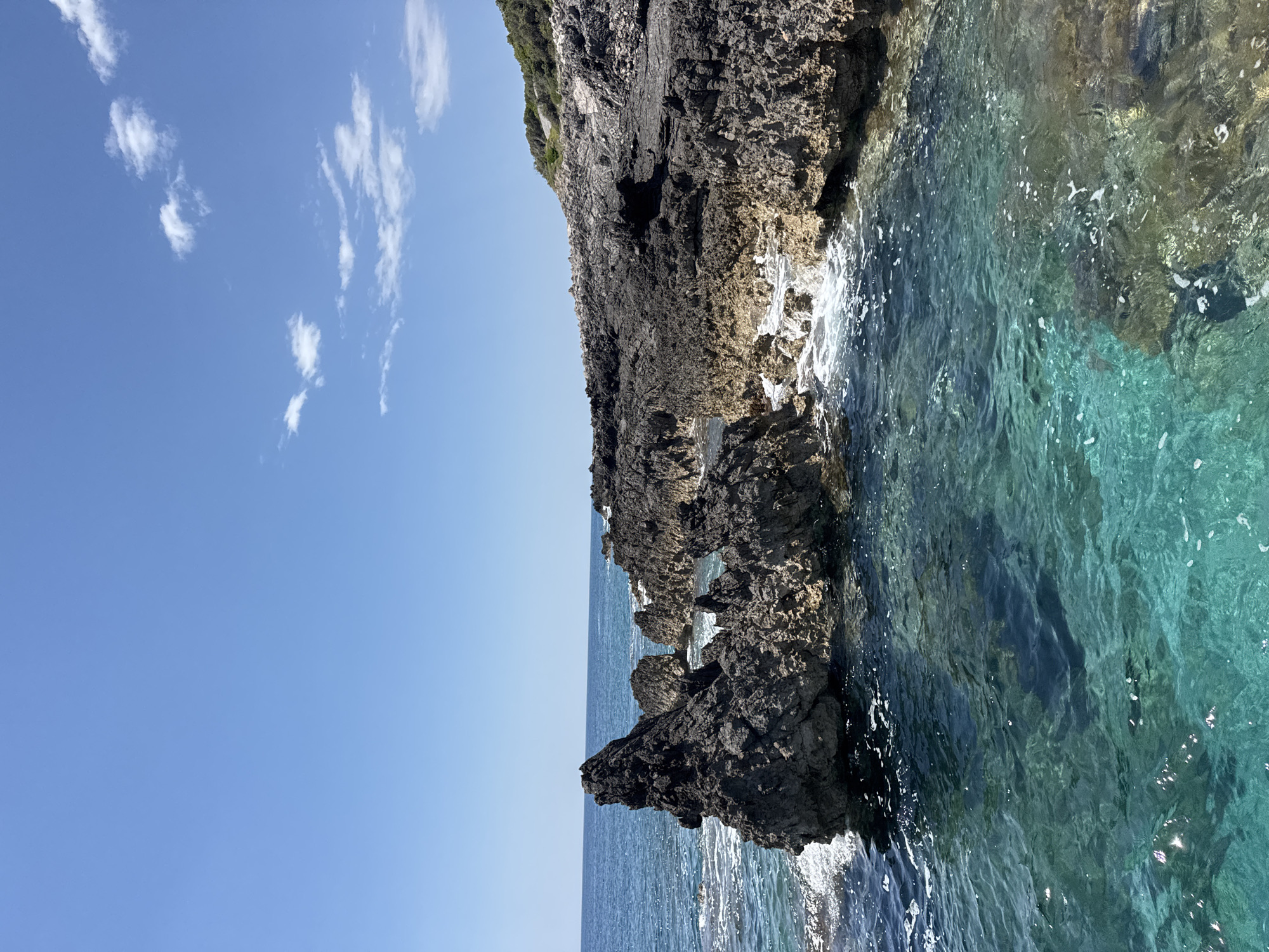 The image shows a rocky shoreline meeting a clear turquoise sea under a bright blue sky with scattered white clouds. The rocks are dark and rugged, with some vegetation visible on the upper portion. The sea is clear enough to see details beneath the surface, and there is evidence of wave action along the rocks. The horizon is visible in the distance. The location is Dubraljevina, Montenegro. There are no people visible in the frame.