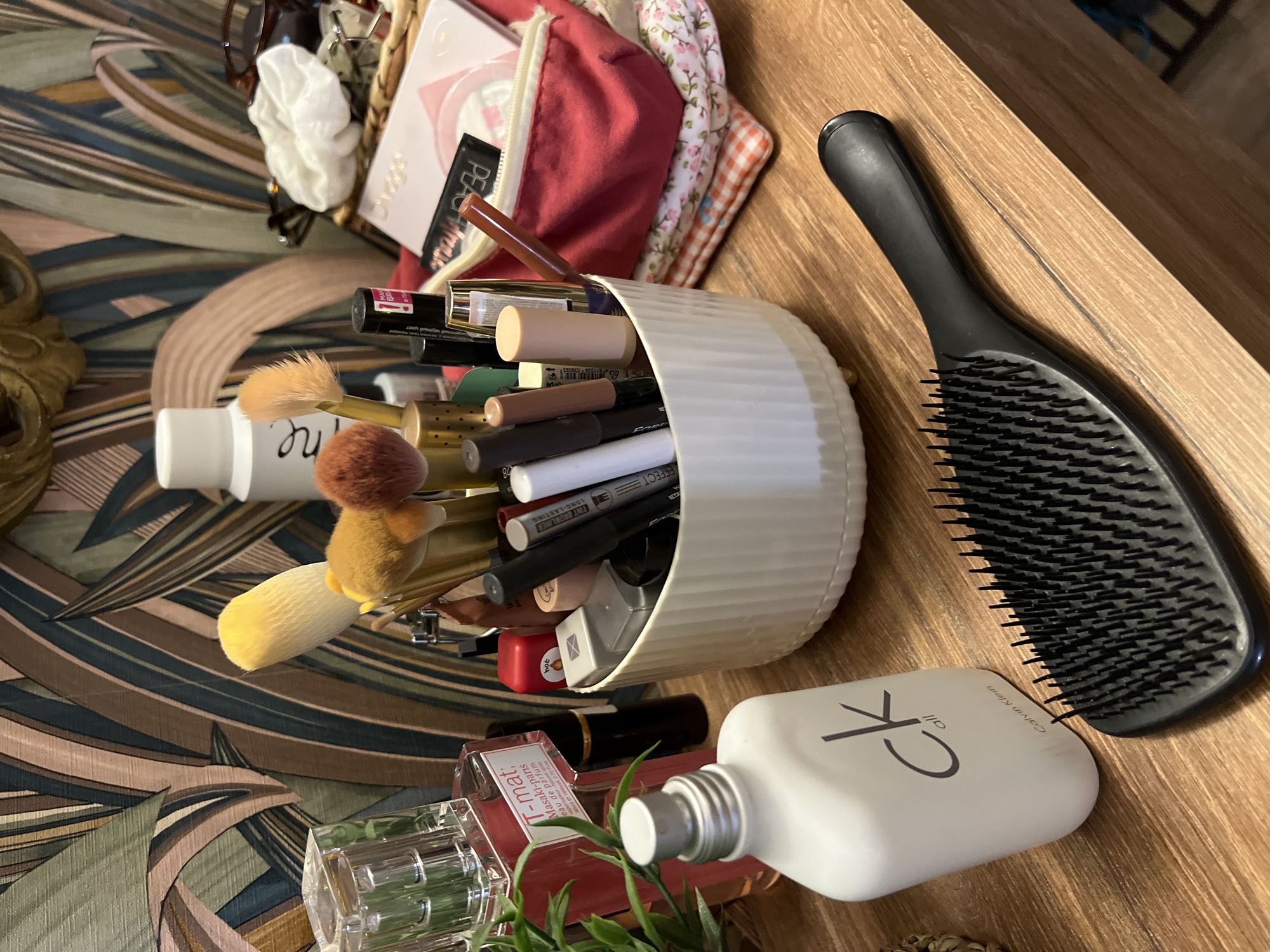 The image depicts a close-up, high-angle shot of a wooden vanity surface cluttered with various cosmetic items and toiletries. Prominently featured in the foreground is a black hairbrush with densely packed bristles and a smooth handle. To its left sits a white, matte bottle of "ck all Calvin Klein" fragrance with its cap on. Behind these items, a white fluted bowl overflows with makeup brushes, eyeliners, lipsticks, and other small cosmetic containers. Further back, a large, clear glass perfume bottle with a pink liquid is visible, adjacent to a potted green plant. To the right, a red makeup bag is stacked on top of floral and checkered fabric. A white bottle with text is also present, along with other indistinct cosmetic products. The background is a wall adorned with a patterned wallpaper featuring abstract, flowing lines in shades of green, brown, pink, and dark blue. The overall scene is a still life of personal grooming products. The image does not contain any visual information that places it in the city of Sinyavino, Russia.