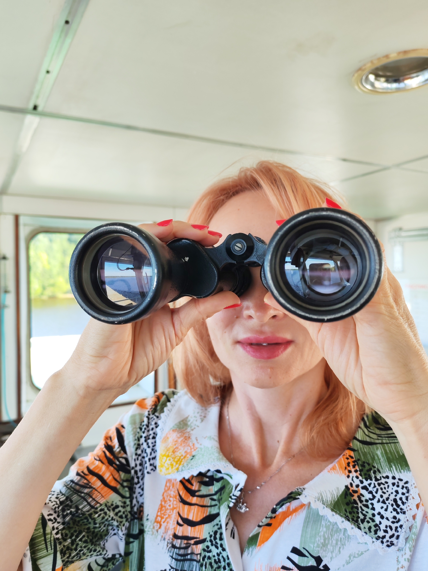 A woman with light-colored hair and bright red nail polish looks through a pair of black binoculars. She is wearing a colorful shirt with a print of green leaves, orange shapes, and black animal spots. A silver necklace with a small charm is visible around her neck. The woman is inside what appears to be a cabin or control room, suggested by the white ceiling with a light fixture and a metal railing. Through the binoculars, a body of water and distant trees are visible, indicating she is likely on a boat or near a waterfront. The lighting suggests it is daytime, and the overall atmosphere is one of observation or anticipation. The background shows a window with a view of the outdoors and part of the interior of the room.