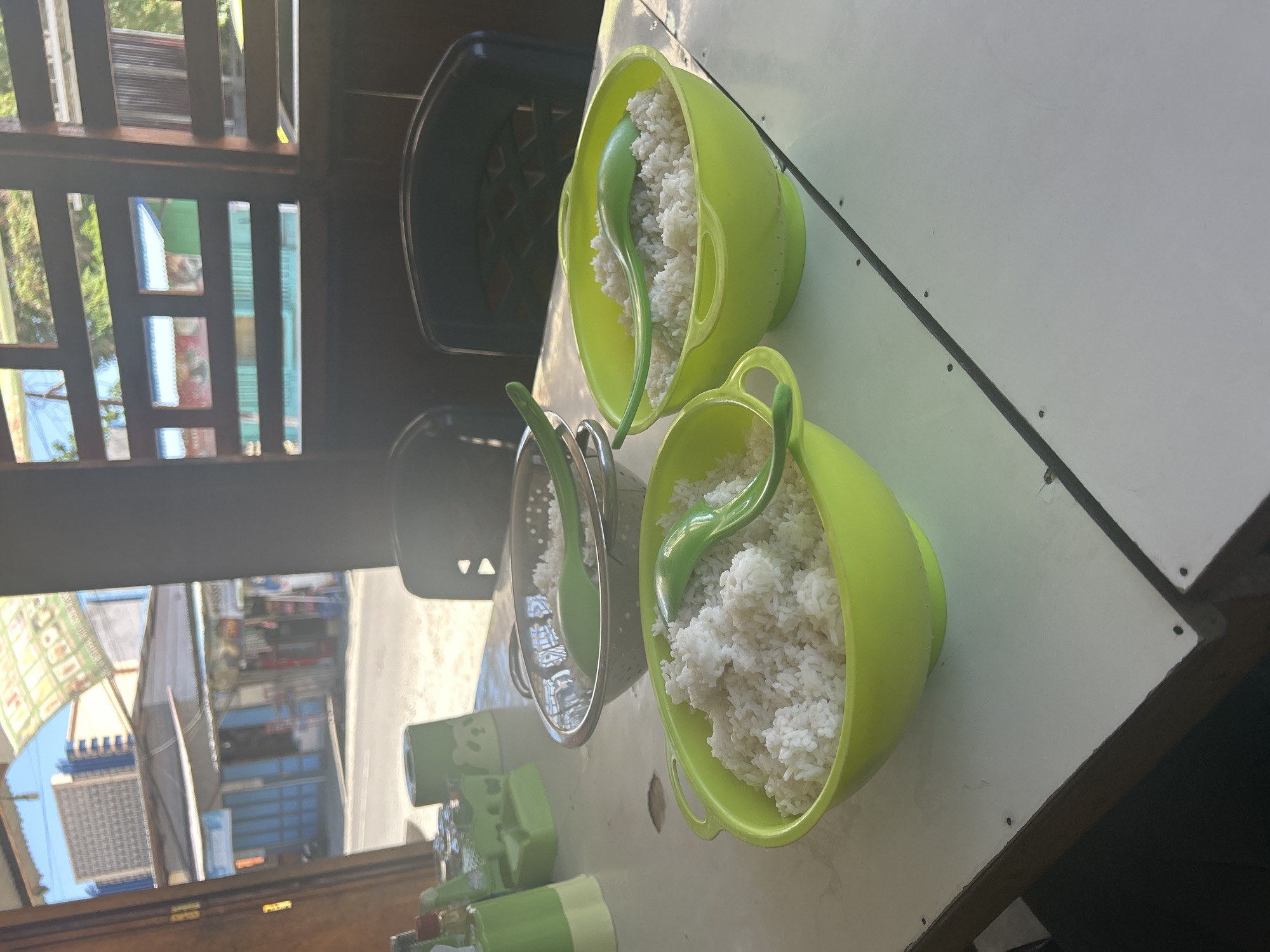 This is a medium shot, slightly tilted, of a white table outdoors, with a blurry background of what appears to be a street scene. The main subjects are three green bowls filled with white rice, each with a matching green serving spoon. The bowls are arranged diagonally across the table. The bowl closest to the viewer is a large, bright green plastic colander-like bowl. To its left, slightly further away and also in focus, is a similar, smaller green bowl. Further to the left and out of focus is a silver metal colander also containing rice.
In the background, through a wooden slatted window or screen, there are glimpses of buildings, greenery, and what looks like shops or stalls. The lighting suggests it is daytime, with bright sunlight casting shadows. The overall impression is that of a casual outdoor dining setting, possibly at a roadside eatery or a home with an open-air eating area. There is no visible text in the image. The scene conveys a simple, everyday meal being prepared or served.