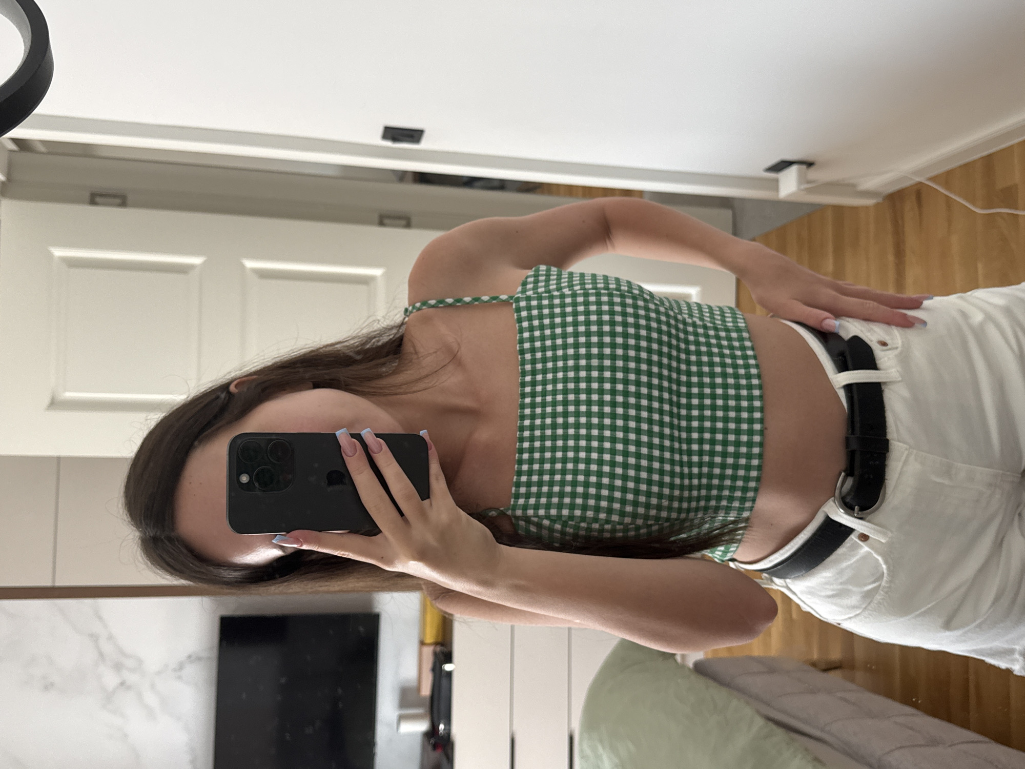 A young woman, wearing a green and white gingham crop top and white denim shorts with a black belt, stands in an indoor setting and takes a mirror selfie with her phone. The background includes a dresser with a television above it, a bed with light green bedding, a wooden floor, and a white door.