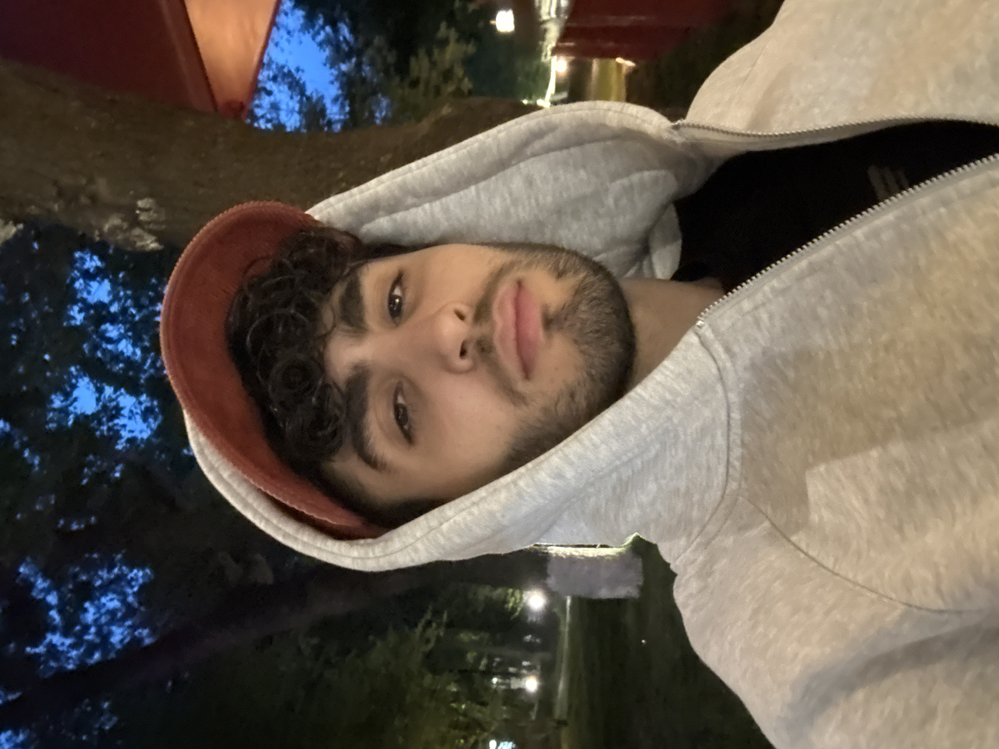 A young man with curly dark hair and a trimmed beard is the primary subject of the image. He's wearing a grey hooded sweatshirt and what appears to be a baseball cap underneath the hood, and the sweatshirt is partially unzipped. The photo seems to be taken at night, possibly outdoors. The background features trees and city lights. The photo could have been taken in Yerevan, Armenia, but without further information, that is an assumption.