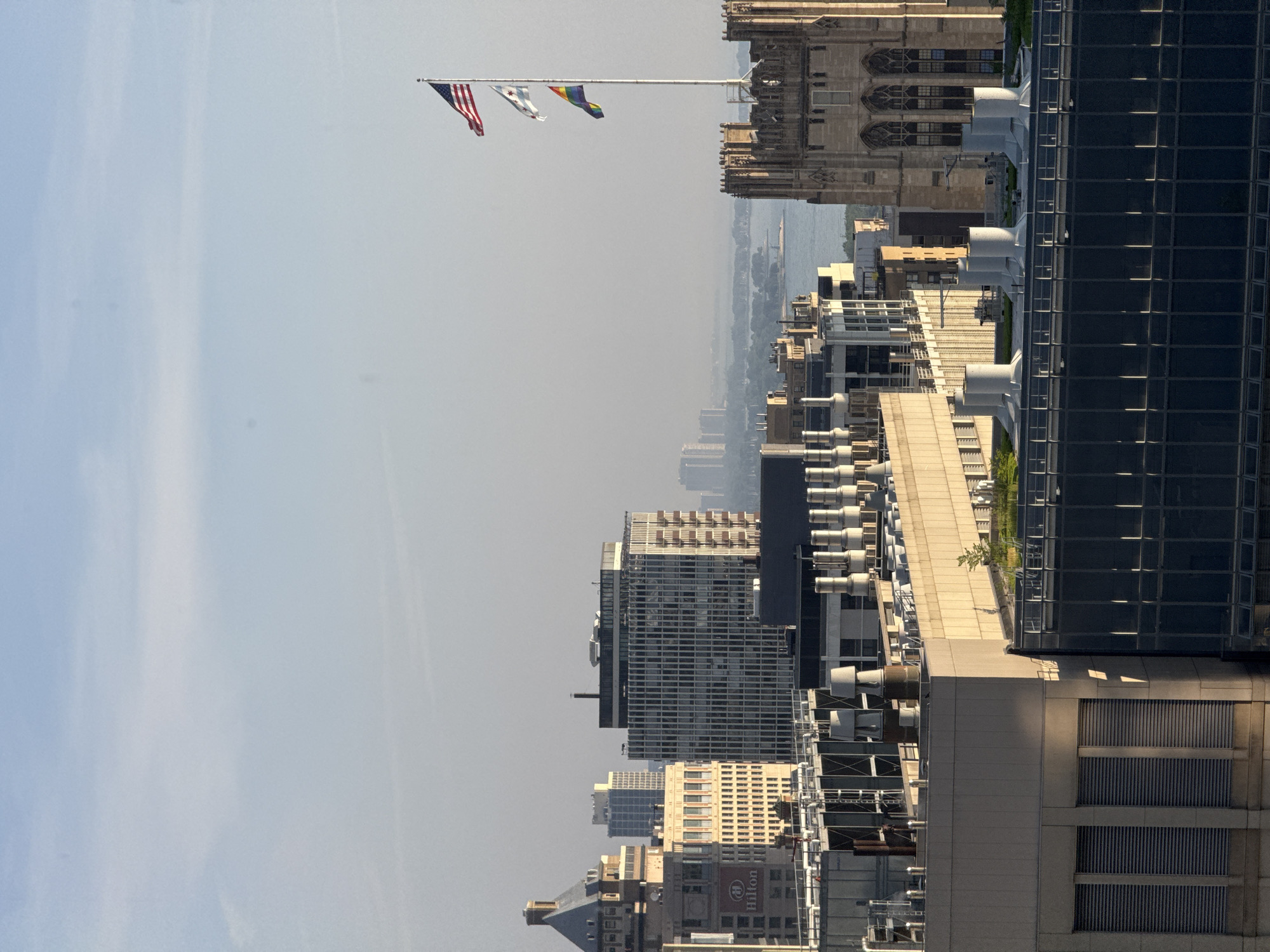 This is a medium-distance, eye-level, outdoor shot of a hazy cityscape in Chicago, Illinois, United States of America. The image is dominated by tall buildings and a pale blue sky. In the foreground, several rooftop HVAC units and architectural details of modern buildings are visible. In the mid-ground, a variety of buildings of different architectural styles are present, including a building with a visible "Hilton" logo. The background features a body of water, likely Lake Michigan, with distant hazy skyscrapers. A flagpole with three flags is prominently displayed on the right side of the image: the United States flag at the top, followed by the Chicago city flag, and a rainbow pride flag at the bottom. The lighting suggests it is either early morning or late afternoon, with long shadows cast on some of the buildings. There are no people or animals visible in the frame.