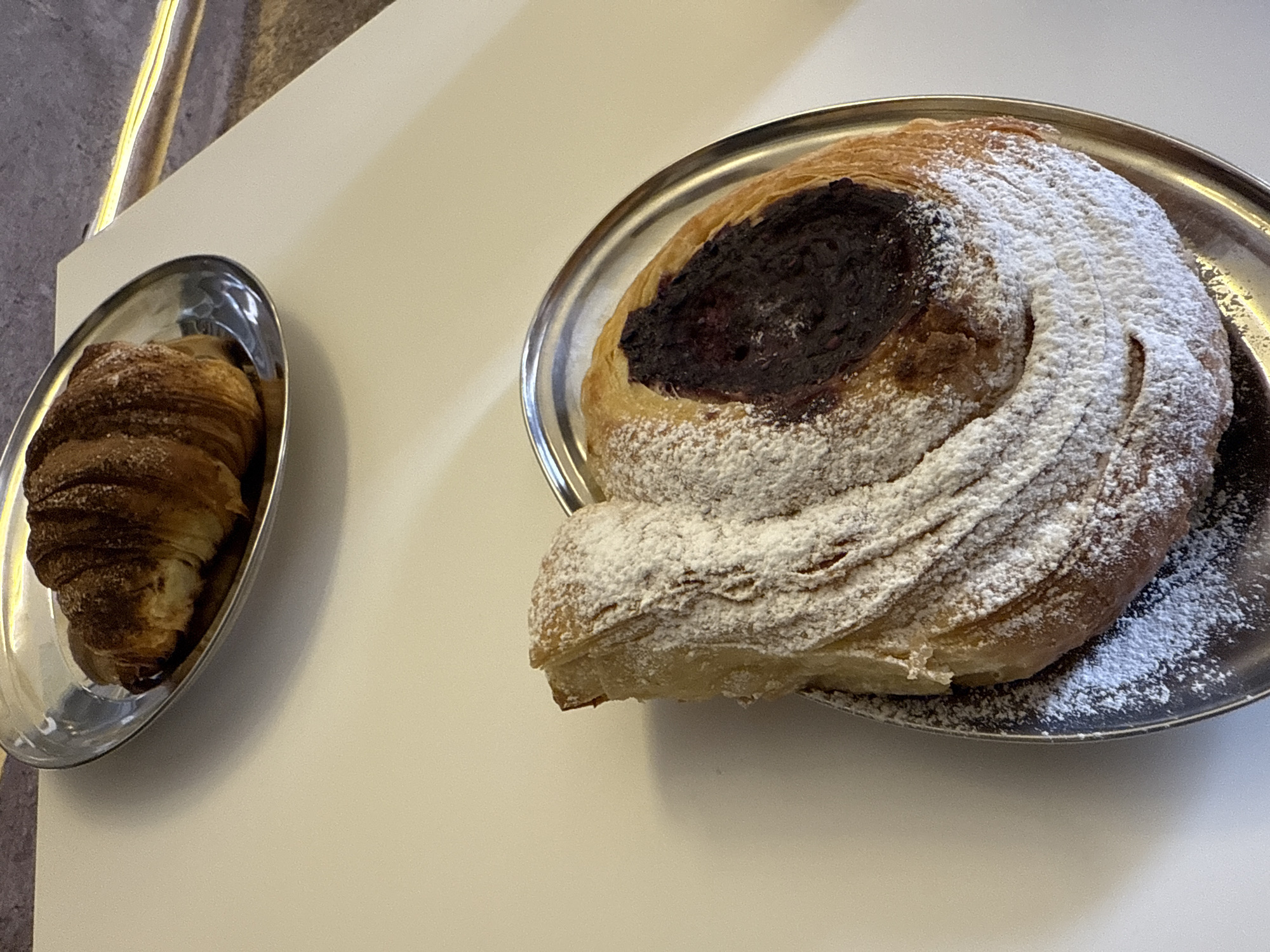 The image displays two pastries served on metallic plates, positioned on a clean, light-colored table or counter. On the left, an oval, shiny silver plate holds a golden-brown croissant, its layered texture visible. To the right, a larger, round silver plate presents a substantial pastry. This larger pastry is generously covered with white powdered sugar, which emphasizes its spiral shape and textured surface. A dark, possibly fruit jam or chocolate filling, appears in the center of the large pastry.
The setting appears to be indoors, possibly a cafe or bakery, with a bright, even light illuminating the scene. A textured, grayish wall is visible in the background on the upper left, alongside a thin, vertical yellow element, possibly a decorative strip or wire. No people are visible, and there is no discernible text. The pastries are the sole subjects, presented for consumption. The context provided is Puebla, Mexico.
