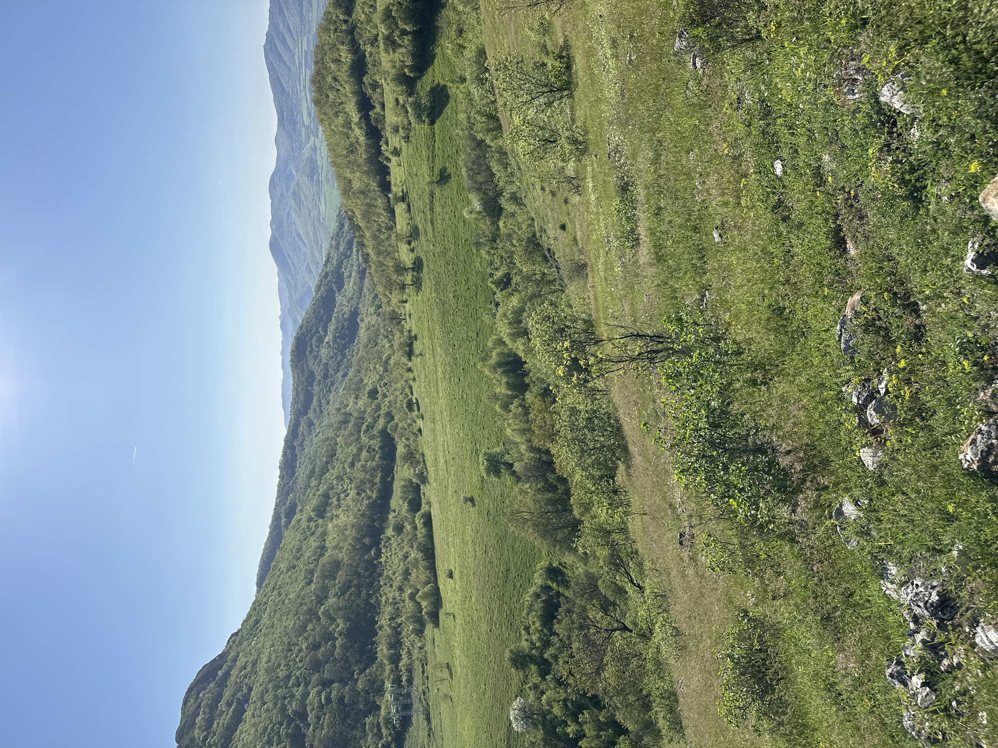The image shows a view of a mountain landscape near the village of Bilka, Bulgaria. The foreground consists of a steep grassy slope with patches of shrubs and exposed rocks. A faint trail or path runs along the slope. The middle ground shows a forested hillside with varying shades of green indicating different types of vegetation. In the background, more distant mountain ranges are visible under a clear blue sky with a bright sun.