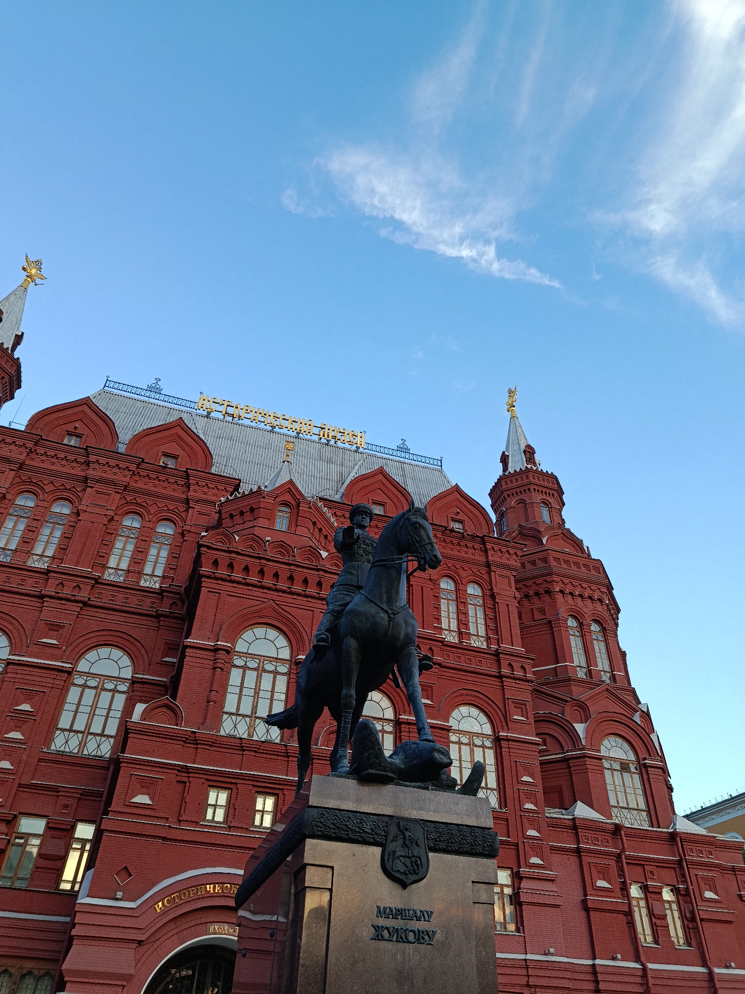 The image captures a grand architectural scene featuring a large, ornate red-brick building and a prominent equestrian statue under a bright blue sky. The building, identified by gold Cyrillic lettering on its roof as "ИСТОРИЧЕСКИЙ МУЗЕЙ" (Historical Museum), showcases a distinctive Russian Revival style with numerous arched windows, intricate brickwork, and grey, patterned roofing. Golden double-headed eagles, symbolic of Russia, adorn the spires and turrets of the building. A partial view of an entrance arch below also displays gold lettering, including "ИСТОРИЧЕСКИЙ" (Historical) and "ВХОД" (Entrance).
In the foreground, a dark bronze equestrian statue depicts a military figure, identified by the inscription "МАРШАЛУ ЖУКОВУ" (To Marshal Zhukov) on its stone base. Marshal Zhukov is shown atop a rearing horse, which appears to be trampling a fallen dragon or serpent-like figure beneath its hooves, symbolizing victory. An emblem is also visible on the statue's plinth.
The sky is a clear, vibrant blue, with a few wispy white clouds trailing across the upper right, indicating fair weather during daylight hours. The light softly illuminates the scene, highlighting the golden details on the museum and the bronze of the statue. The composition, from a low angle, emphasizes the monumentality of both the building and the statue. This iconic view points to the location being Moscow, specifically the State Historical Museum and the Marshal Zhukov statue near Red Square. The image conveys a sense of history, national pride, and architectural grandeur.