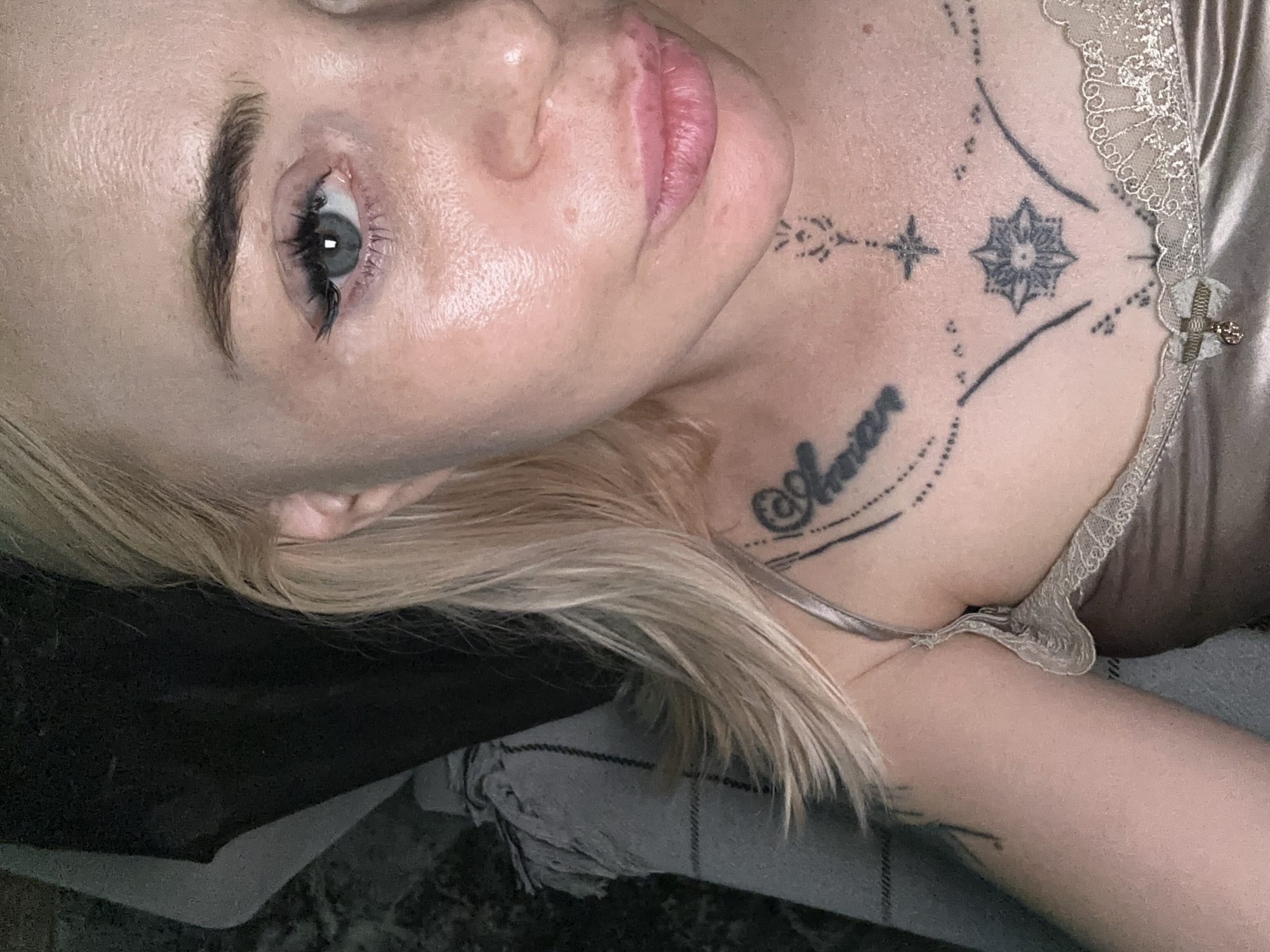 A close-up portrait depicts an individual, appearing to be female, with blonde hair and fair skin, gazing directly at the camera. The right eye is visible, featuring a blue-green iris, dark eyeliner, and prominent false eyelashes. The forehead and right cheek exhibit a glossy sheen. The lips are full and tinted pink. The individual wears a light beige or champagne-colored satin garment with a lace-trimmed neckline, accented by a small bow and a metallic charm at the center. Multiple tattoos are present on the chest and upper arm: a script tattoo on the viewer's left chest, an arrangement of geometric and ornamental designs including a starburst and a cross-like symbol on the central chest, and a smaller stylized pattern on the lower neck. The background consists of a dark, indistinct area on the upper left and a light-colored, patterned textile, possibly a blanket, on the lower left. The lighting suggests an indoor environment. The specific location in the city of Unknown, Unknown is not identifiable.