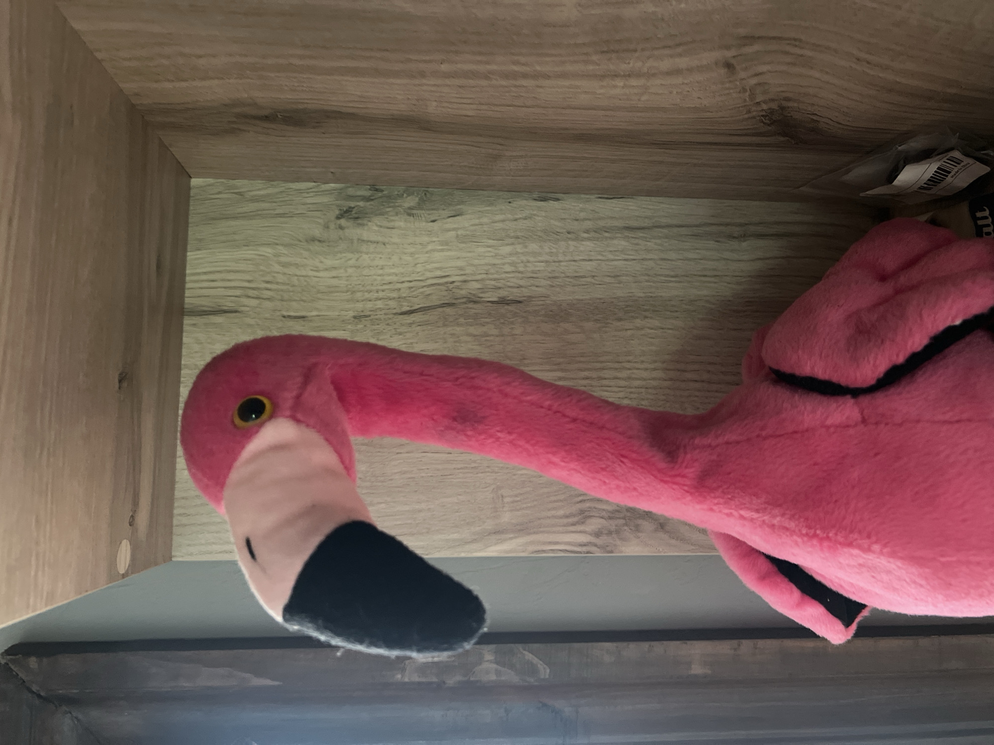 A plush pink flamingo is positioned inside a wooden shelf. The flamingo's head, with a black beak and yellow eye, is prominently displayed, angled towards the left. Its long, curved neck extends from a fluffy pink body. The shelf has a light wood grain texture, and the overall lighting suggests an indoor setting.