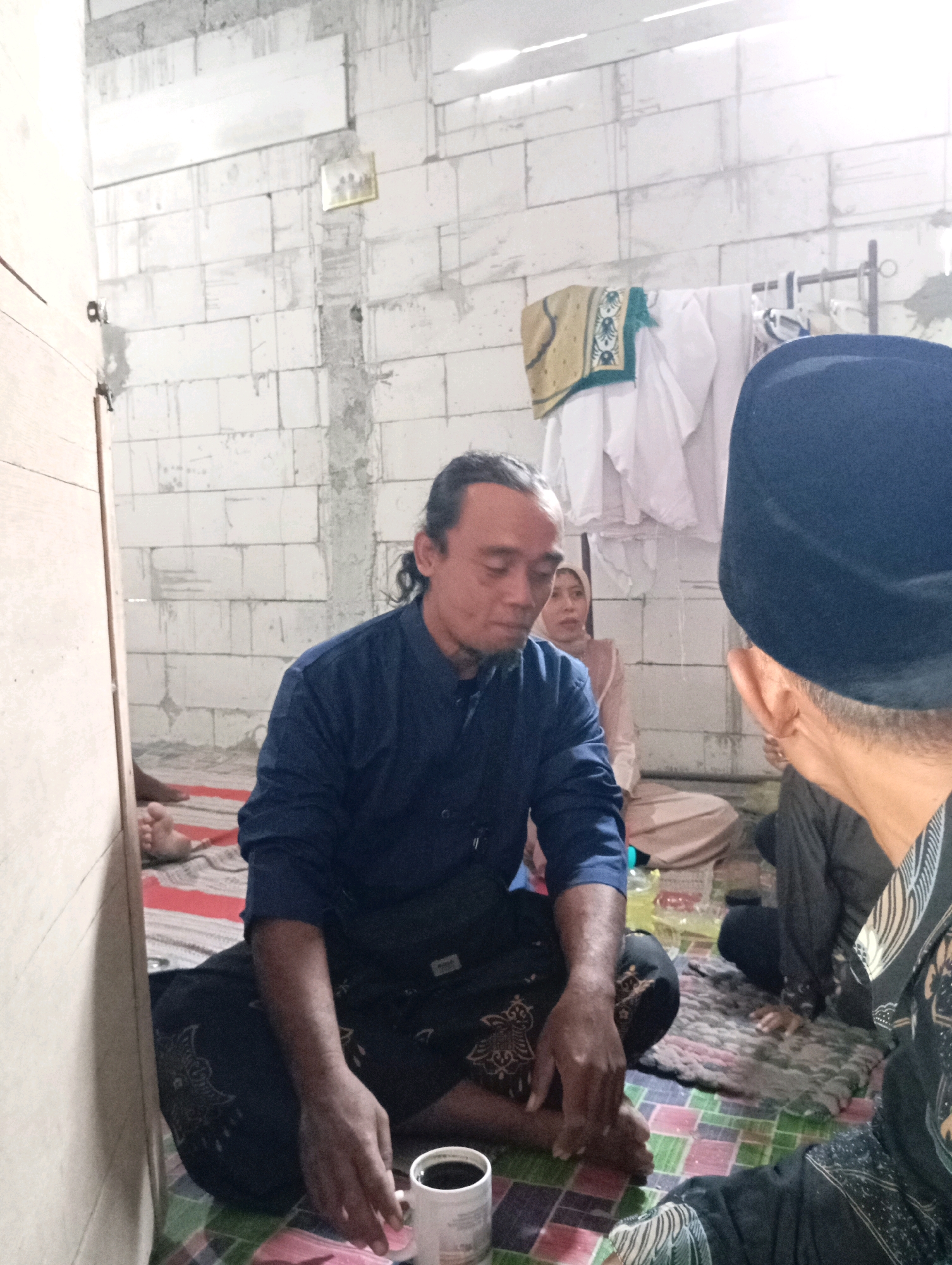 In the city of Ngampel, Indonesia, inside a room with unfinished cinder block walls, a group of individuals are gathered. The room appears to be a residential space, evidenced by the presence of a clothes rack with garments hanging on it. In the foreground, a man with dark, tied-back hair wears a navy blue long-sleeved shirt and dark patterned pants, sitting on a mat. He is holding a white mug filled with a dark liquid. To his right, another man, wearing a dark peci (cap), is partially visible from behind. In the background, a woman in a light-colored hijab and traditional clothing is seated. The floor is covered with various mats, including one with a checkered pattern. The overall impression is of an indoor gathering, possibly a social or religious event.