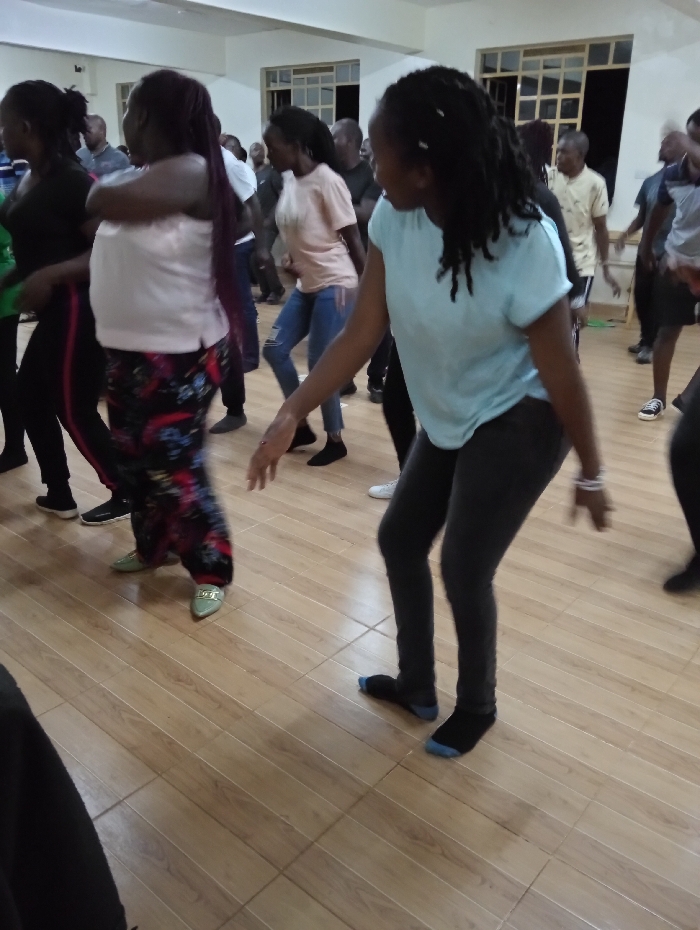 This media file captures a group of people, primarily adults, engaged in an energetic indoor activity that appears to be dancing or group exercise. The scene is set in a spacious room, possibly a community hall or a large studio, featuring a light-toned wooden floor with tile-like divisions and pale walls. Several windows with white frames are visible in the background, allowing for natural light, though artificial lighting may also be in use.
In the foreground on the right, a woman with dreadlocks, wearing a light blue t-shirt, dark leggings, and black and blue socks, is actively moving with her body turned to the left. In the mid-ground on the left, another woman with long dark hair, dressed in a light pink top, colorful patterned pants, and light green pointed flats, is also in motion with her arms crossed. Further to her left, a woman in a black top and black leggings with a red stripe is visible.
Numerous other individuals, both men and women, are participating in the background, some appearing blurred due to movement, indicating a dynamic group activity. Most participants are dressed in casual attire such as t-shirts, jeans, and leggings. Some are seen wearing socks directly on the wooden floor, while others have shoes. The atmosphere suggests a lively and communal event. There is no visible text in the image. The location context indicates this event is taking place in Riara Ridge, Kenya.