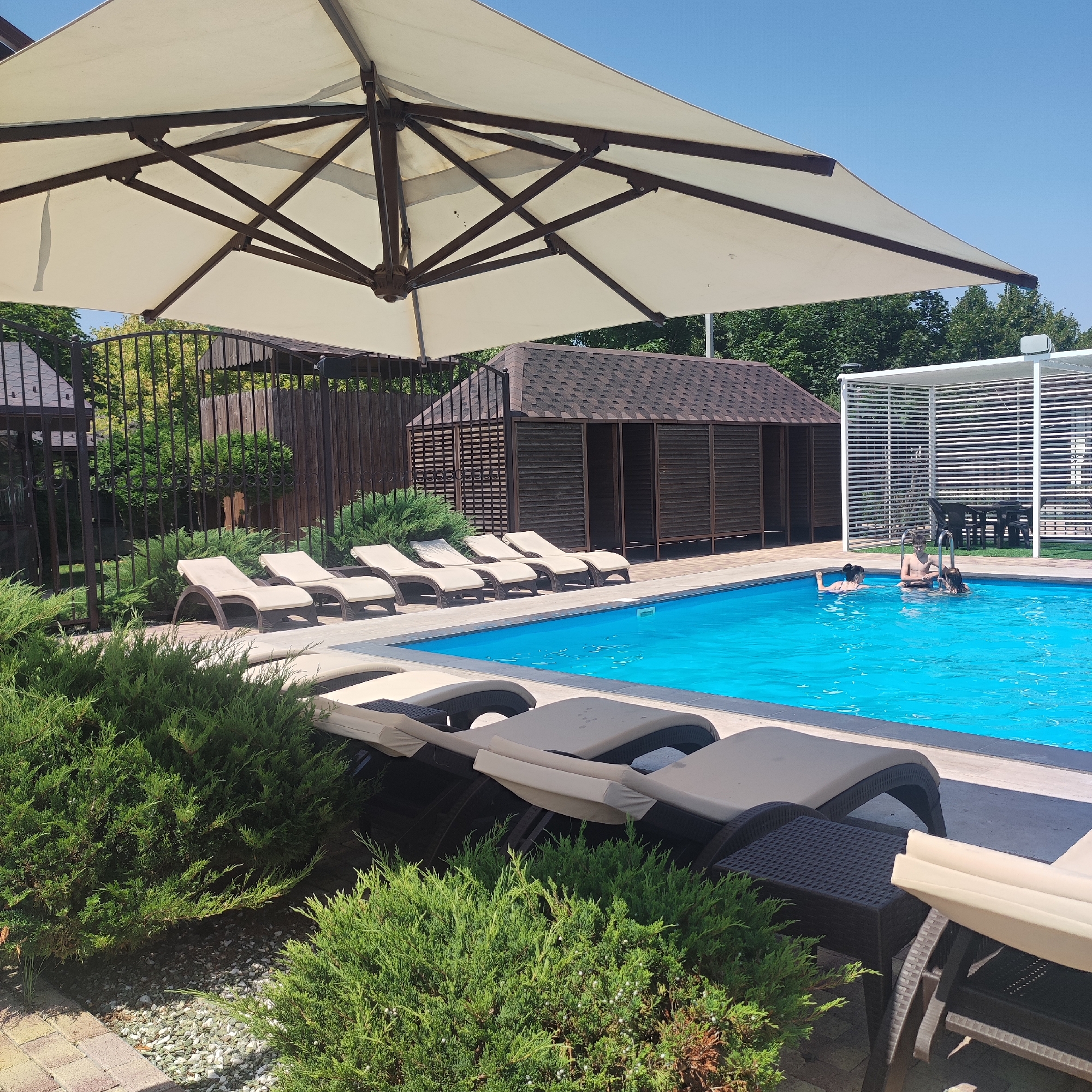 The image depicts a sunny day at an outdoor swimming pool complex. In the foreground, several beige lounge chairs are arranged on a paved area, partially obscured by lush green bushes. A large, cream-colored umbrella provides shade over some of the lounge chairs. The swimming pool itself is a vibrant blue, with three people swimming: one person on the left appearing to be swimming, another in the center holding onto the pool ladder, and a third person on the right also in the water. Adjacent to the pool is a modern white structure with horizontal slats, and beyond that, a dark-colored gazebo with a sloped, shingled roof. Further in the background, a wooden fence encloses the area, with trees and clear blue sky visible above. The overall scene conveys a relaxed atmosphere of a recreational outdoor space.