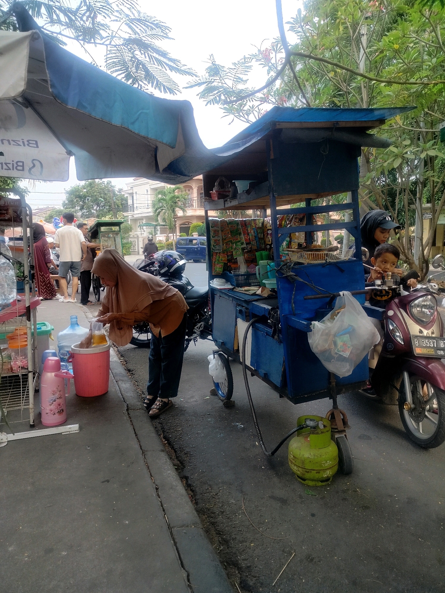 In East Jakarta, Indonesia, a street vendor is preparing a drink at a mobile cart. The vendor, a woman wearing a tan hijab and brown tunic, is seen pouring a yellow liquid into a plastic bag, holding a large pink cooler in her left hand. Beside her, on a metal rack, are several large water bottles, a pink thermos, and other containers. Behind her, a blue cart, likely a food or drink stall, is attached to a motorcycle. A woman in a black hijab and a young boy are seated on the motorcycle, facing the cart. The scene unfolds on a paved street with a curb. Trees and buildings are visible in the background, suggesting an urban environment. A green gas cylinder is positioned on the ground near the cart.