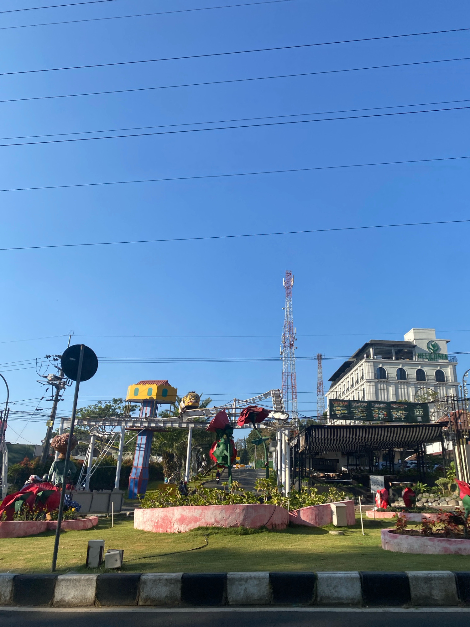 The image captures a street-level view of a commercial area under a clear blue sky. In the foreground, a paved road with a black and white striped curb is visible. Beyond the curb, a neatly manicured lawn is bordered by a pink concrete planter filled with greenery and red floral accents. Various colorful, cartoonish sculptures, some resembling figures and others abstract shapes, are dotted across the lawn. These sculptures are situated on and around an elevated metal structure, suggesting an amusement or recreational area.
To the left, a utility pole with visible wires and a circular black sign stands prominently. In the middle ground, a tall, red and white telecommunications tower rises against the sky. Behind the sculptures and tower, a modern, multi-story white building with arched windows and a green logo on its facade is situated. A black and white striped awning covers an outdoor seating area in front of this building. A banner displaying country names and flags, along with "12K", is also visible on the building's facade. The overall scene depicts a vibrant and somewhat eclectic urban environment on a bright, sunny day.