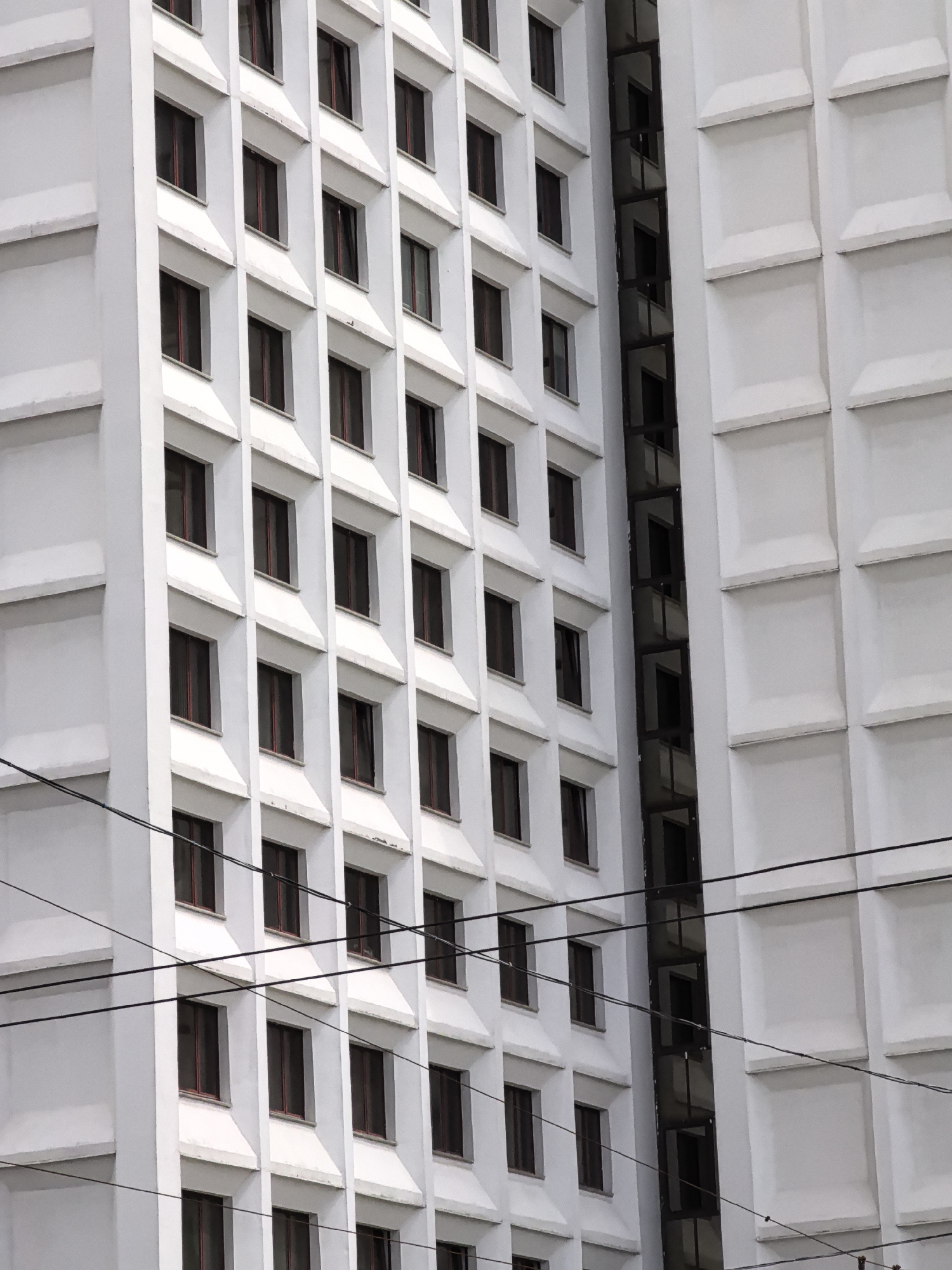 This is a close-up, eye-level shot of a modern, white, multi-story building with many windows. The building's facade is highly geometric, with repeating square windows set within recessed frames. The windows themselves appear dark, suggesting they are either tinted or the interiors are unlit. The building has a sleek, minimalist design. To the right of the main facade, another section of the building is visible, featuring larger, square textured panels. Several dark electrical wires traverse the foreground, running horizontally across the frame, indicating the presence of an urban environment. The lighting is overcast, casting a soft, diffused light on the structure.