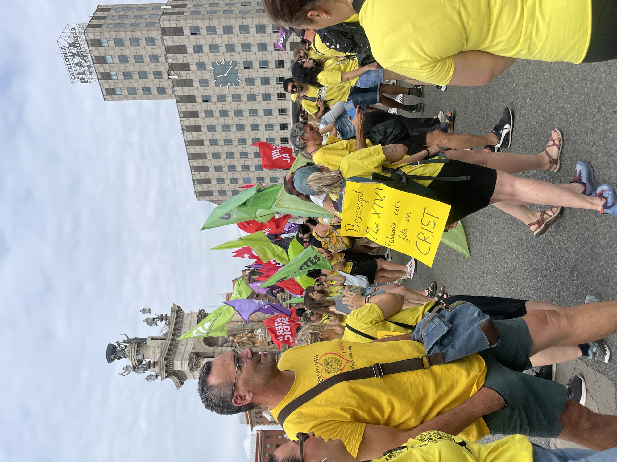 The image shows a crowd of people, many wearing yellow shirts, gathered in a city square. A large, ornate monument with statues is visible in the background, as well as a tall building with the words "CATALONIA PLAZA HOTEL" on top. The building also features a prominent clock face. Several people are holding flags of various colors, including green, purple, and red. One person in the foreground is holding a yellow sign with handwritten text in Catalan. The overall atmosphere appears to be that of a public gathering or protest. This scene is consistent with Plaça de Catalunya in Barcelona, Spain, a central hub for public events and demonstrations.