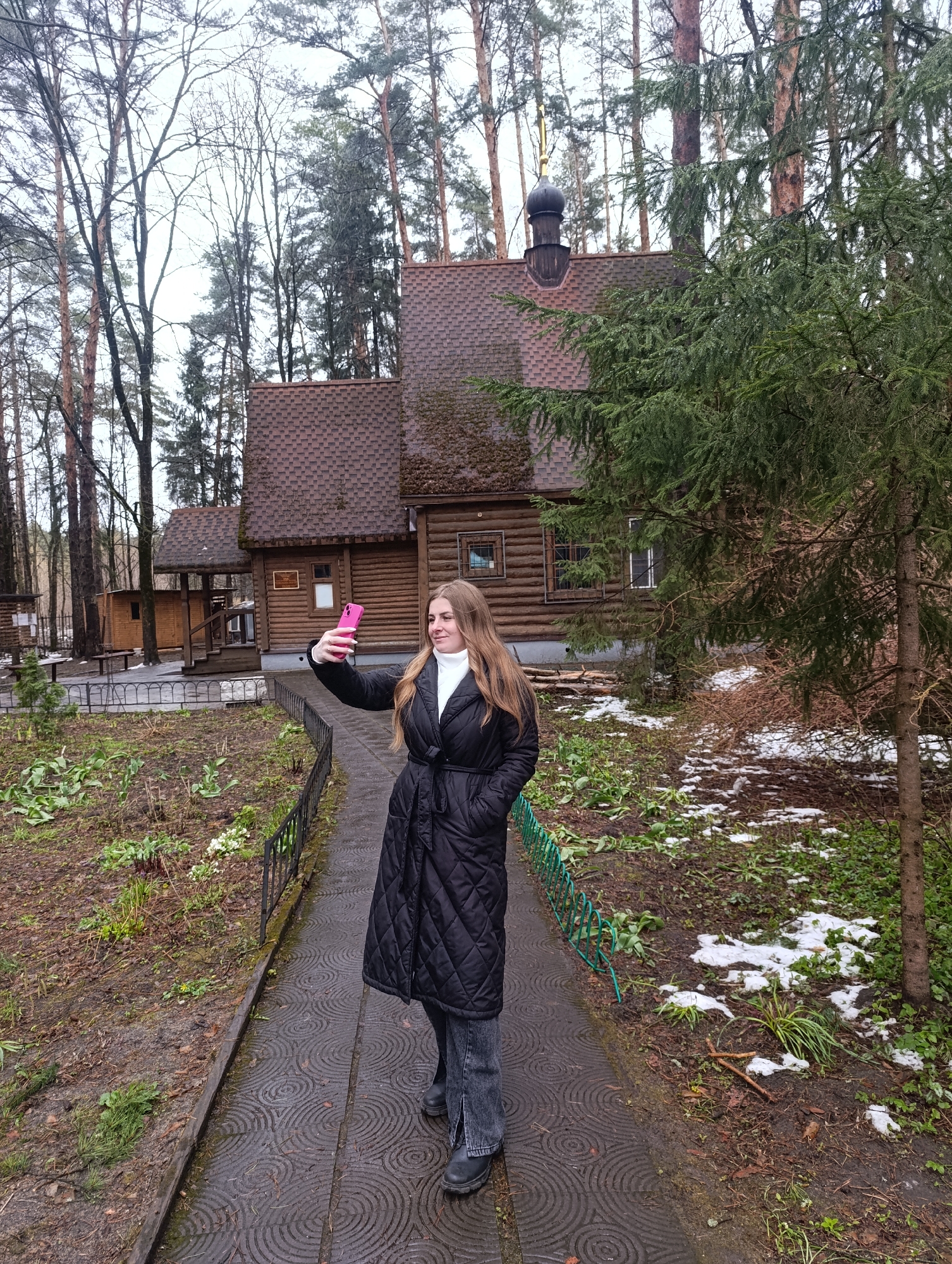 A young woman is captured in a selfie pose, holding a bright pink smartphone, on a paved pathway in what appears to be the grounds of a wooden chapel or church. She wears a black quilted long coat over a white turtleneck and dark pants, with black boots. Her long hair is light brown.
The scene is set outdoors, likely in late winter or early spring, indicated by patches of melting snow on the ground, emerging green sprouts, and some bare deciduous trees interspersed with evergreen pines. The sky is overcast, casting a soft, diffused light.
Behind the woman stands a rustic wooden building, resembling a chapel, with a shingled brown roof and a dark, onion-shaped dome topped with a golden cross. The building features log walls and several small windows. To its left, another smaller wooden structure is visible. The path the woman stands on has a distinctive pattern of concentric circles. A low metal fence, partly green, lines the pathway, separating it from the surrounding ground.
The overall setting suggests a tranquil, natural environment, possibly a spiritual retreat or a historical site in Chernogolovka, Russia. A small white sign with text in an indecipherable script is visible on the side of the main wooden building.