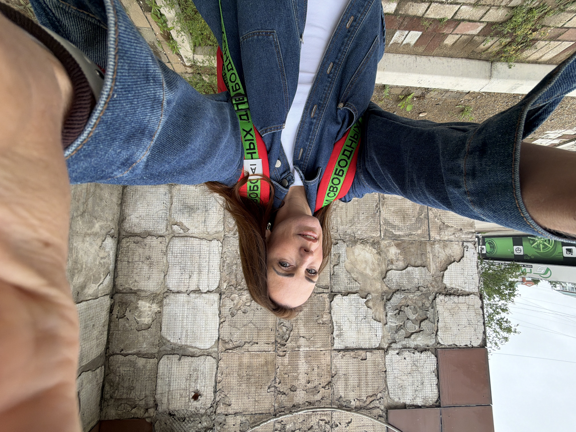An adult Caucasian woman with long brown hair is taking a selfie outdoors in front of a distressed, tiled wall. She is wearing a blue denim jacket over a white top and a red backpack with green straps that have black text. Her arms are extended, holding the camera, and her right hand is visible in the foreground, partially obscuring the image. The background includes a section of a building with brown and white tiles, some of which are peeling and damaged. A street or sidewalk with brick paving and a concrete curb is visible below the wall, with some sparse green vegetation. In the distance, a green building with signage is visible, suggesting an urban environment. This appears to be a street scene in Birobidzhan, Russia, given the context.