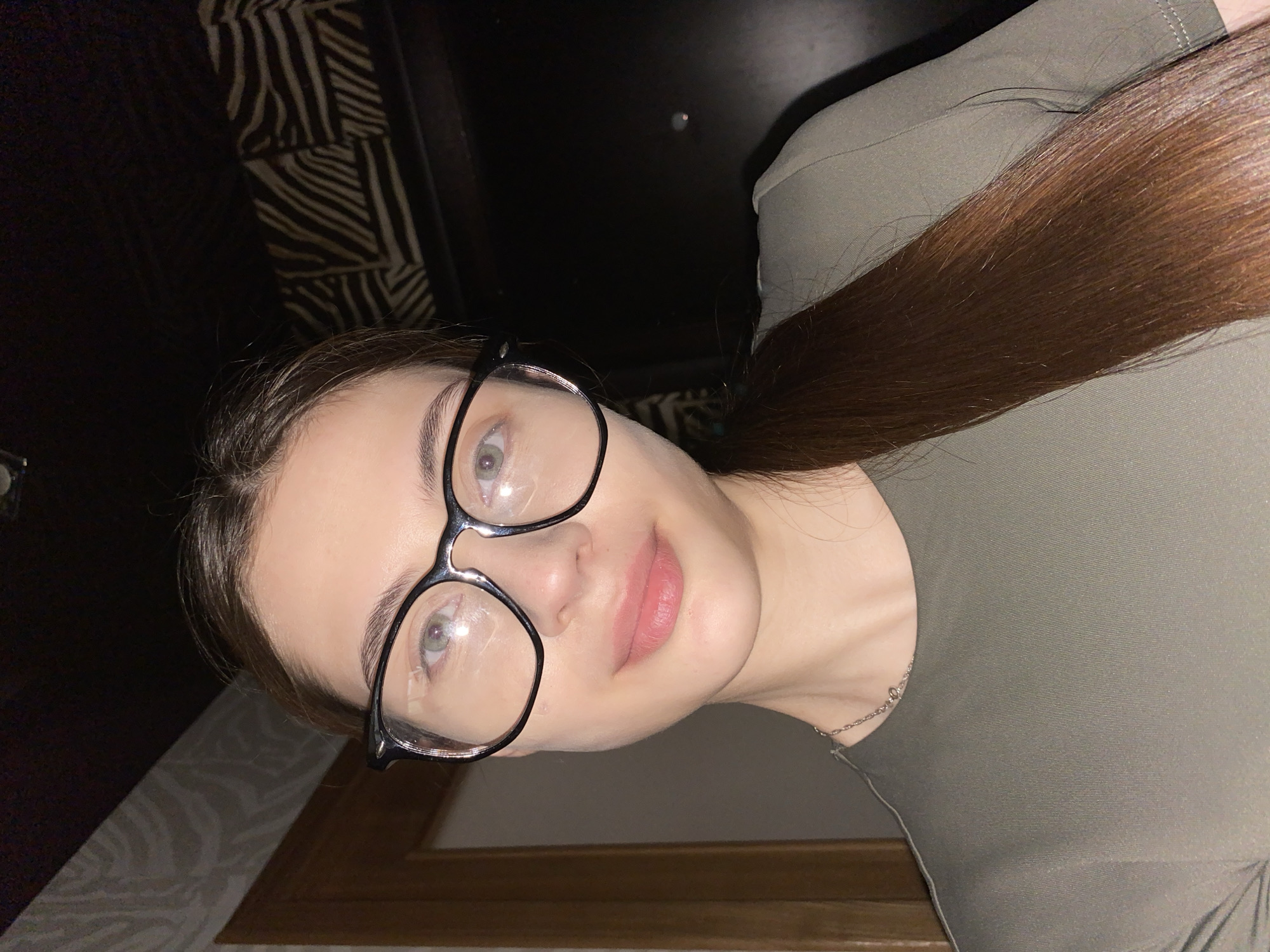A young woman is featured in a close-up indoor portrait, potentially taken in Votkinsk, Russia. She is positioned with her head slightly tilted, looking directly at the camera with a calm and gentle expression. She wears dark-framed eyeglasses, behind which her light-colored eyes are visible, with reflections from an unseen light source on the lenses. Her lips are slightly parted.
She has long, straight, dark brown hair that flows over her right shoulder, extending down the side of her torso. She is wearing a simple olive green or khaki-colored top or dress, and a delicate silver-colored chain or necklace is visible around her neck.
The background is dimly lit and largely dark. On the upper left, there is a dark surface with a distinct patterned texture, resembling a zebra or striped print, possibly belonging to furniture or wallpaper. Below her face, a lighter wall or surface is visible, accented by a dark wooden frame or trim at the bottom edge. The lighting appears to be artificial, suggesting it could be day or night. No text is visible in the image.