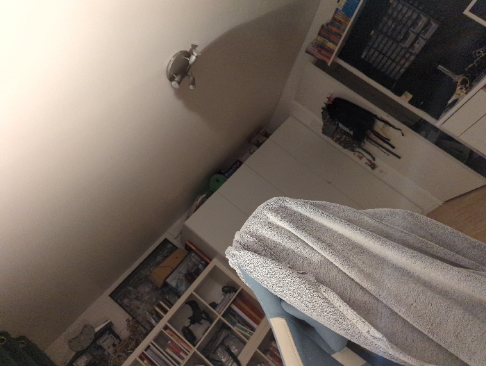 The image shows a white towel, a light gray blanket, and a white door. There are also bookshelves on either side of the door with books, a backpack hanging on hooks, and a shelf with a variety of objects. The ceiling is white with a light fixture in the middle. The location is a bedroom in Lagny-sur-Marne, France, at coordinates 48.879, 2.705. The photo was taken at an upward angle.